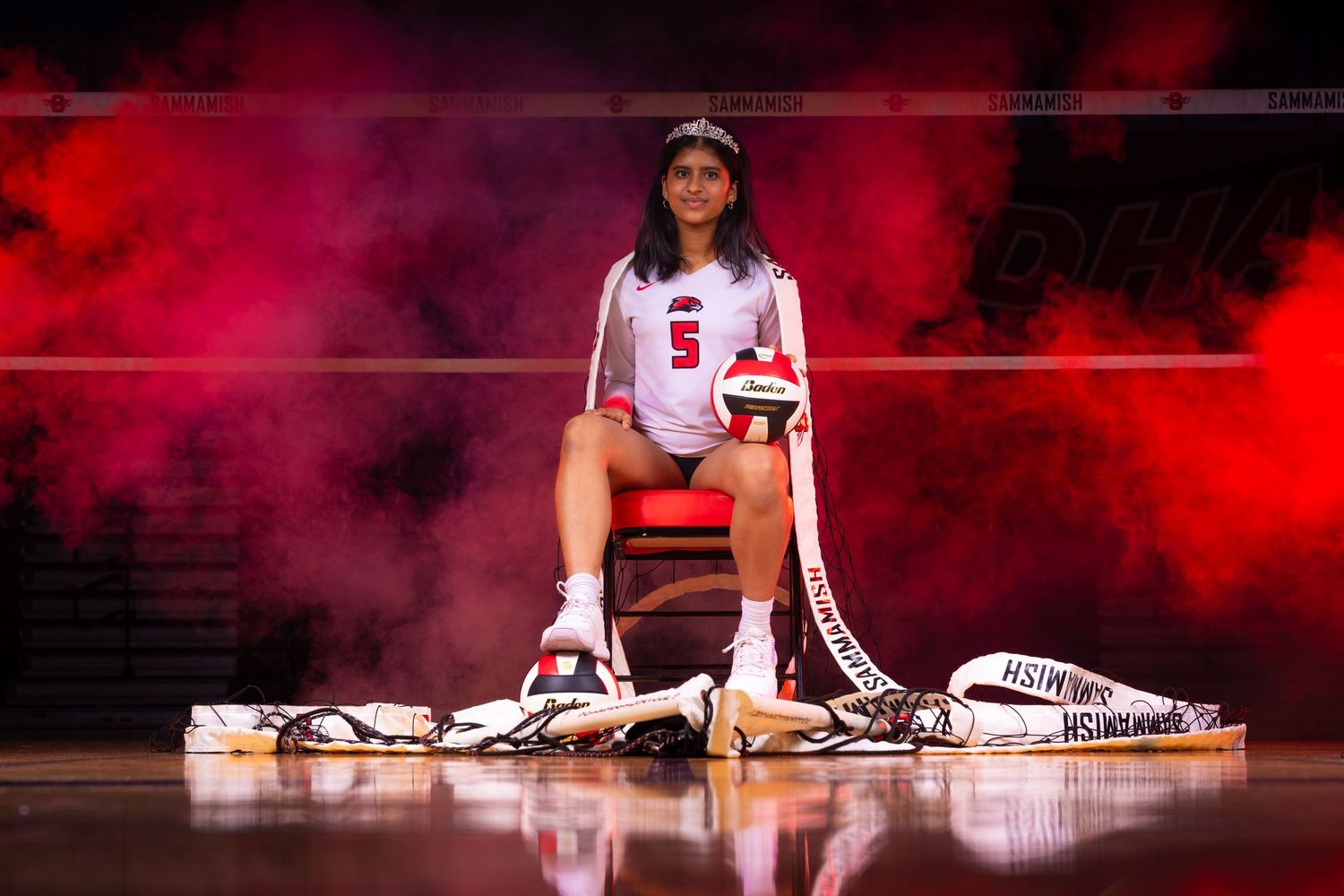 Volleyball player in USA uniform poses dramatically with sports equipment against red smoky backdrop.