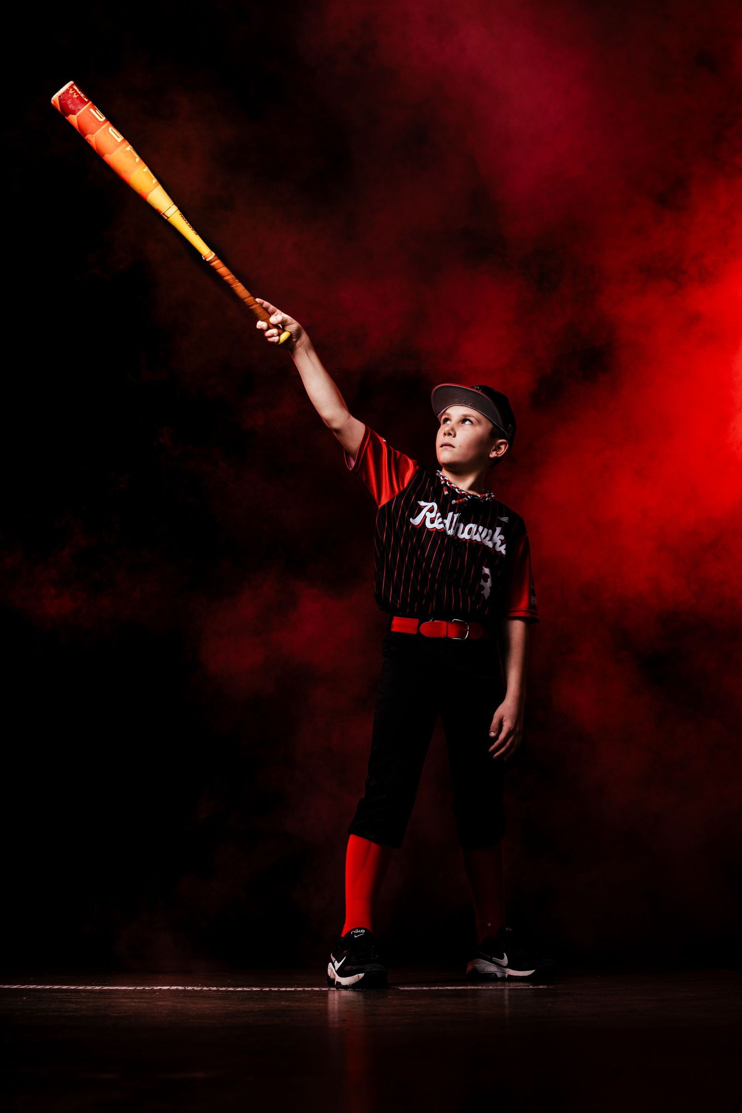 A performer in dark clothing strikes dramatic poses with a baseball bat against a moody red and black backdrop.