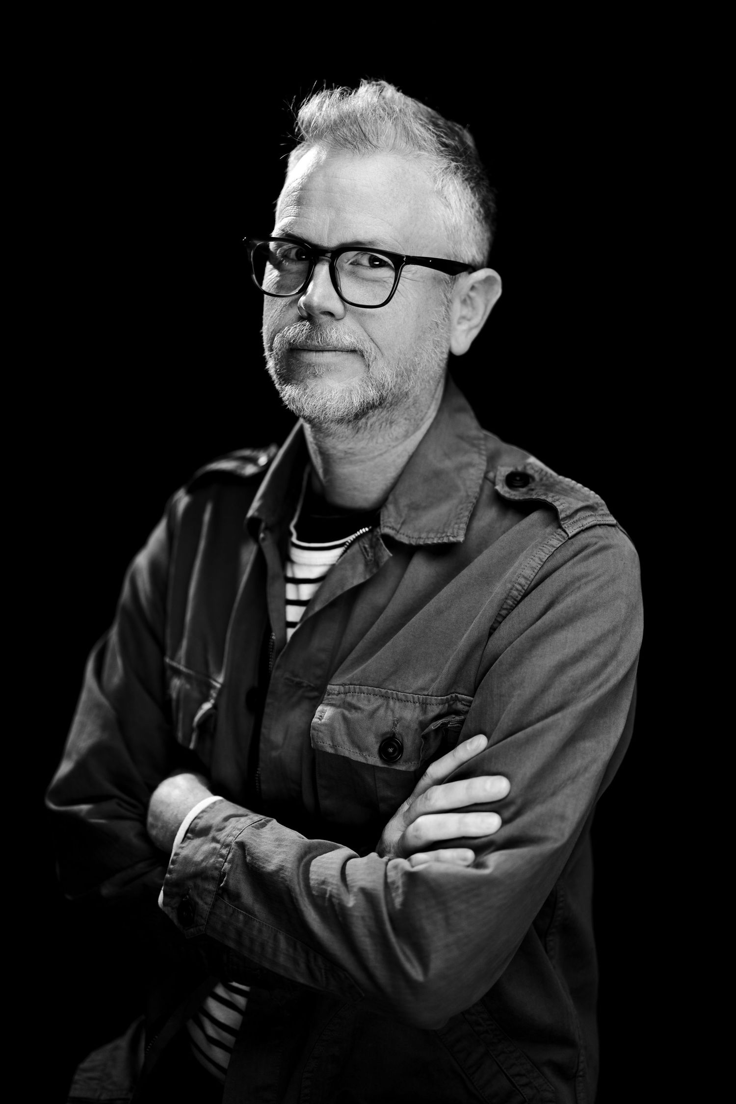 Black and white portrait of person wearing glasses, striped shirt and utility jacket with arms crossed against dark background.