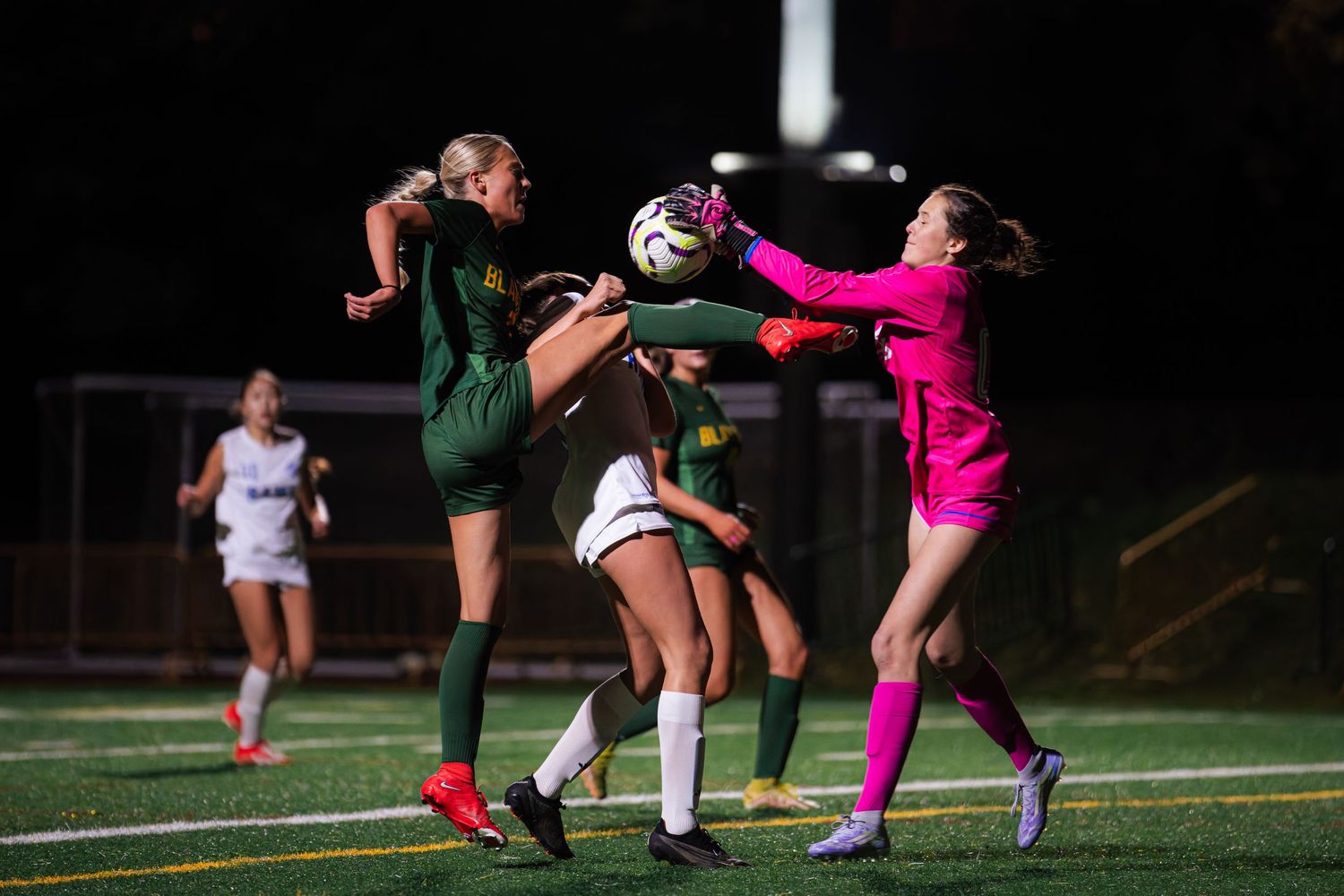 Intense soccer match action shows players competing for the ball during a nighttime game on an illuminated field.
