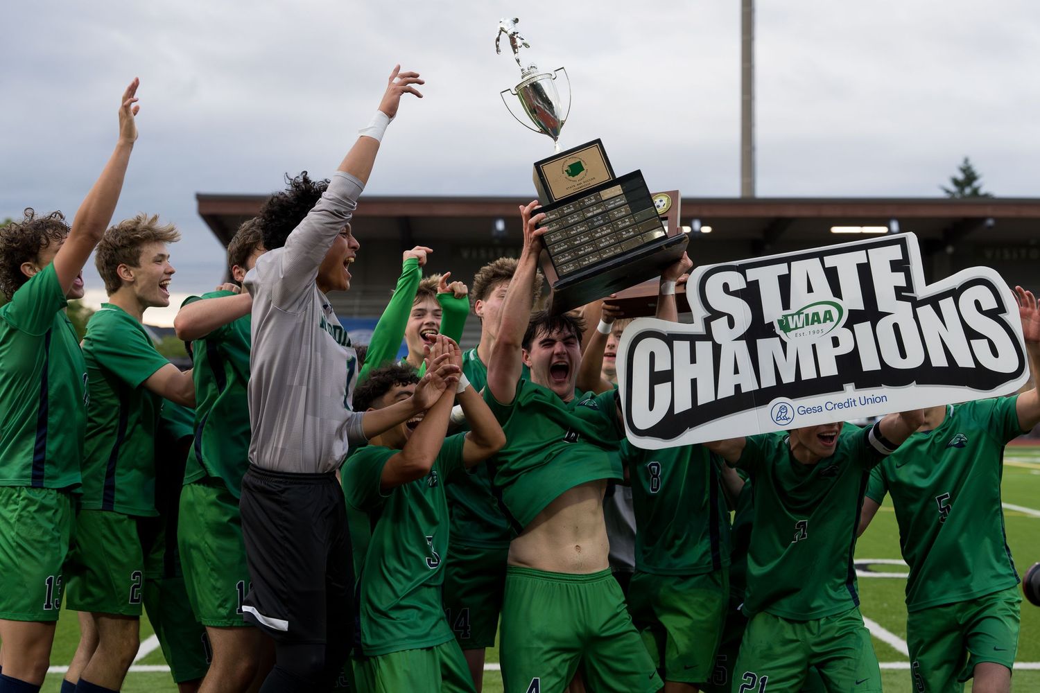 Championship team in green uniforms celebrates with trophy on field.