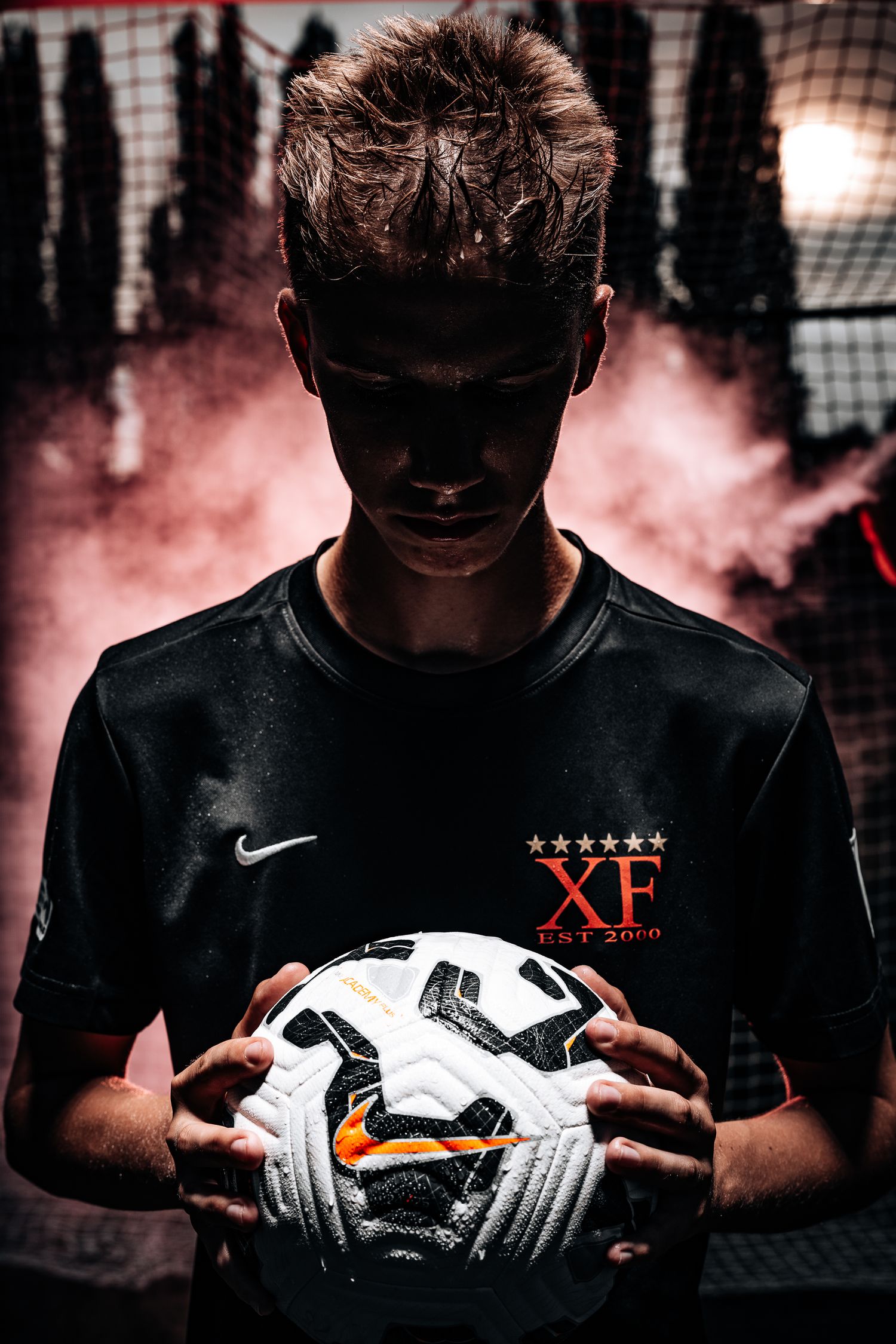Soccer goalkeeper wearing Nike gear holds white ball against dramatic pink smoke background and netting.