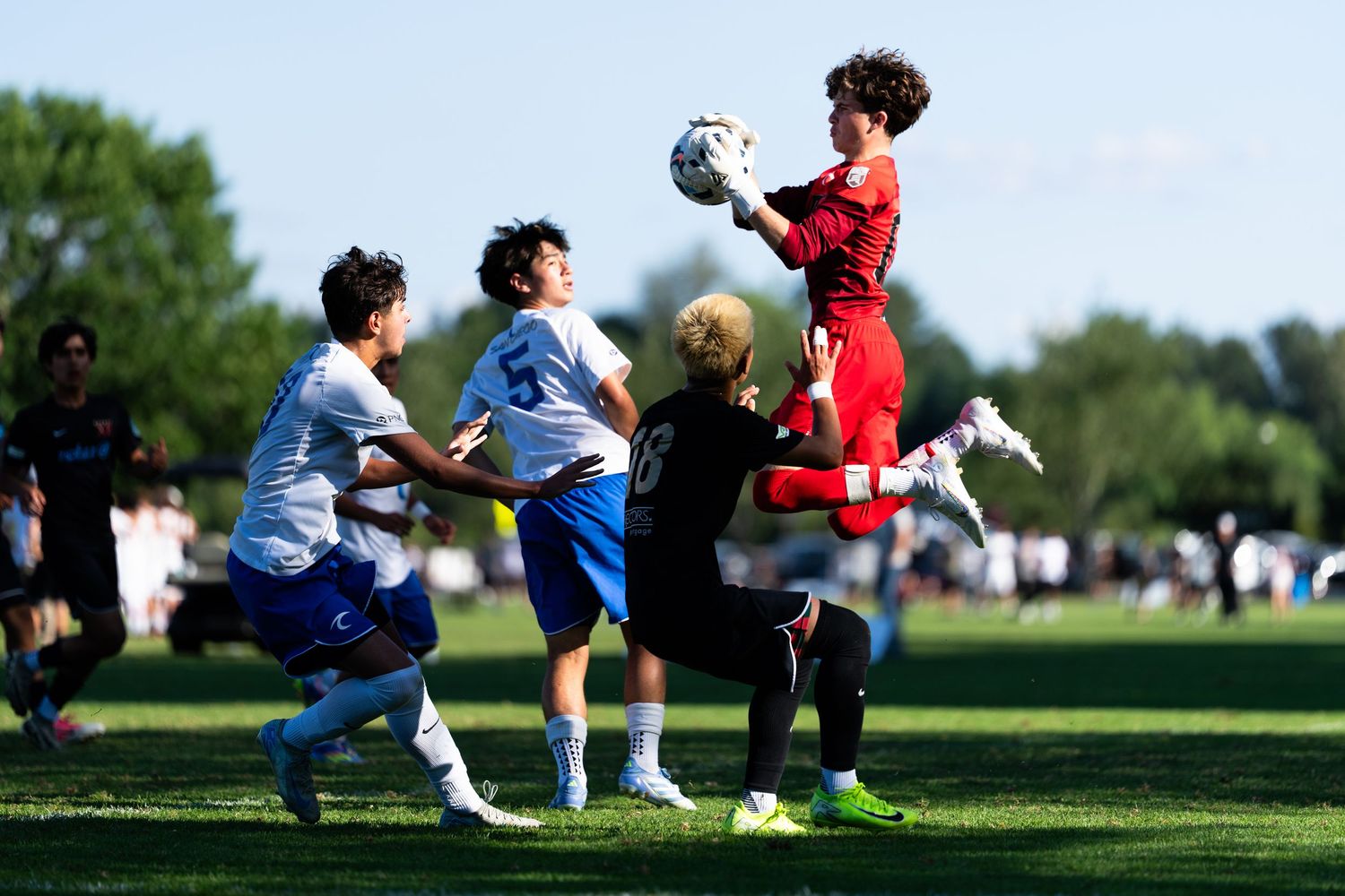 Youth soccer players compete for the ball as a goalkeeper in red makes a leaping save during an outdoor match.