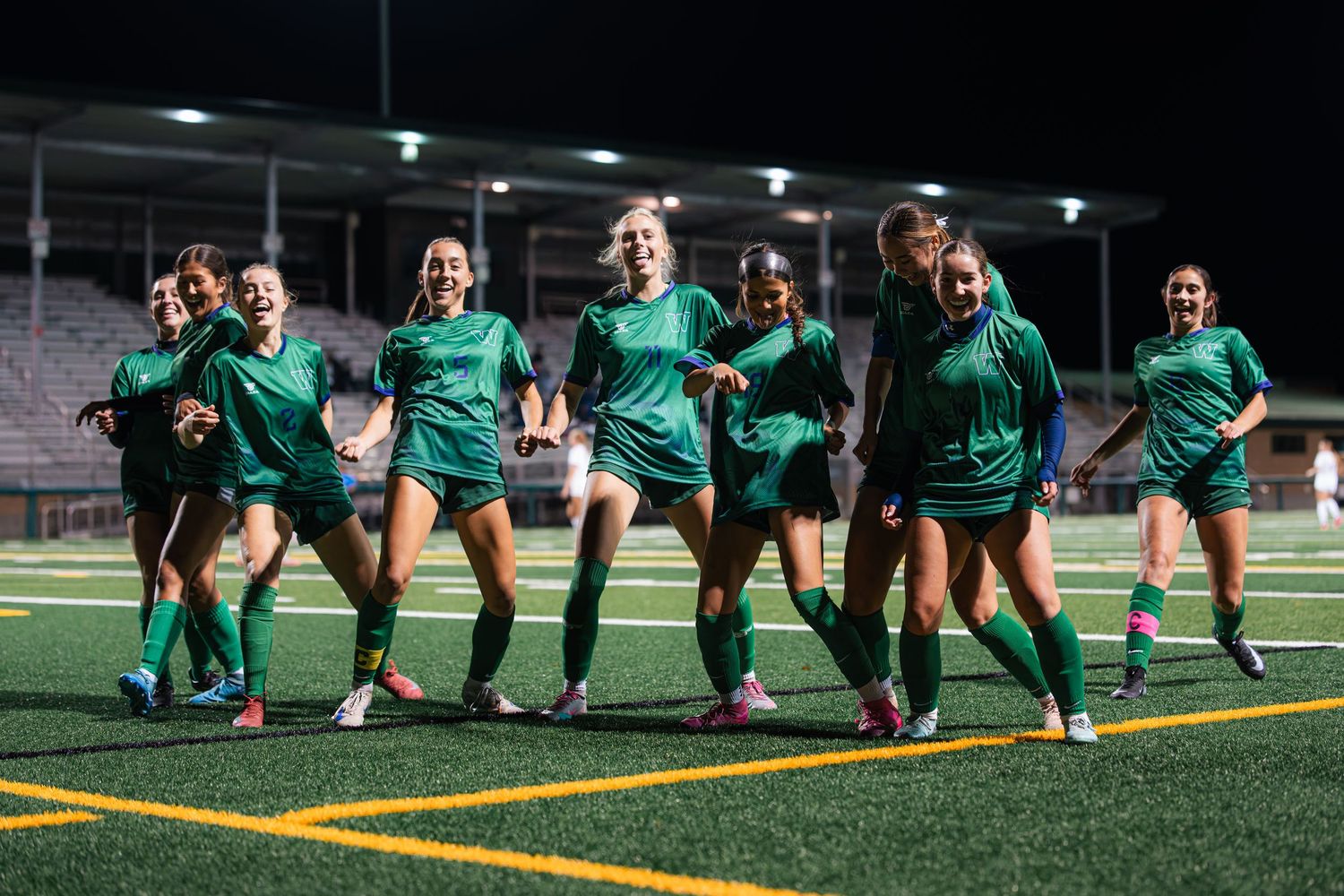 Soccer team in green jerseys performs synchronized stretching exercises on a turf field during practice session.