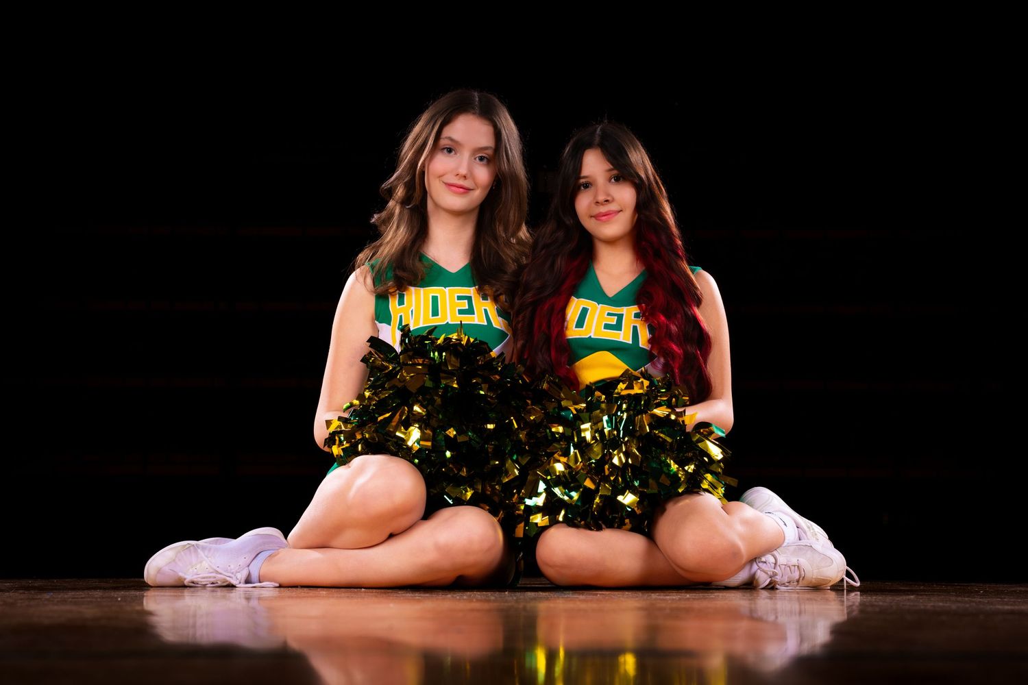 Cheerleaders in green and yellow uniforms pose together during a performance routine on a dark stage.