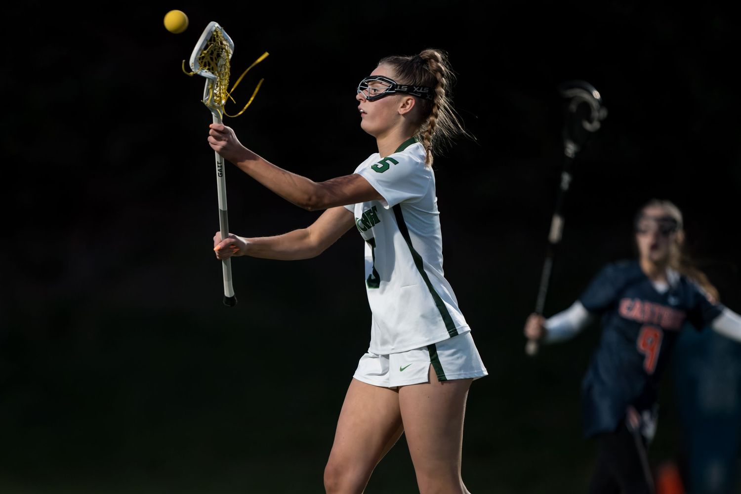 A lacrosse player in a light blue uniform catches the ball with their stick during a nighttime game.