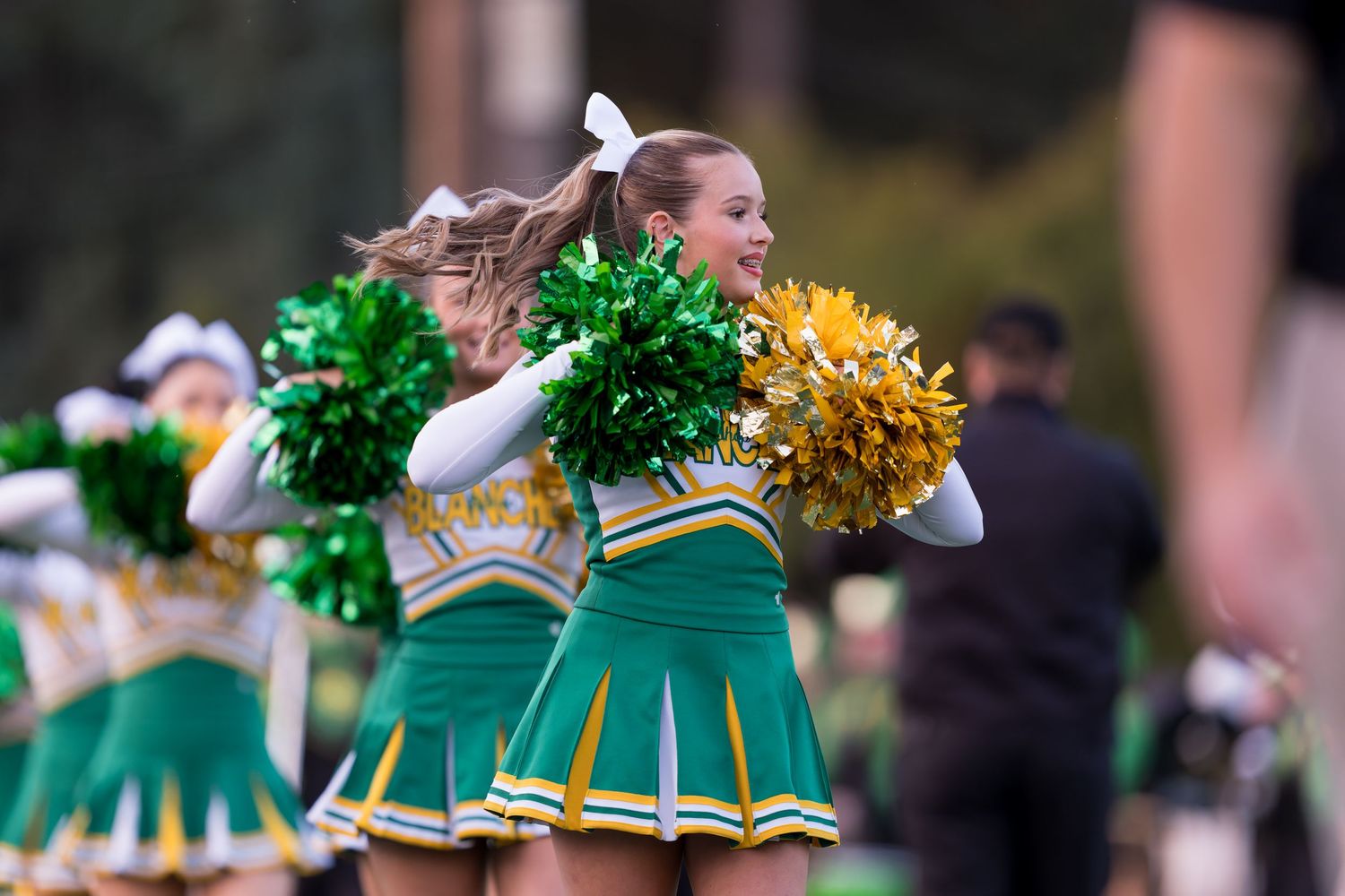 Cheerleader in green and gold uniform performs with pom poms during outdoor sporting event.