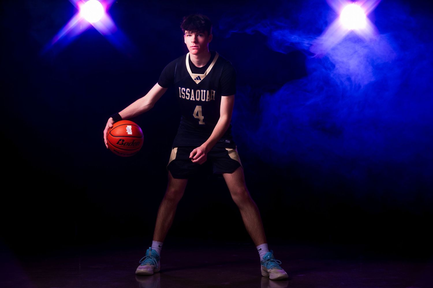 Basketball player in dark uniform dribbles low while making defensive stance against blue background lighting.