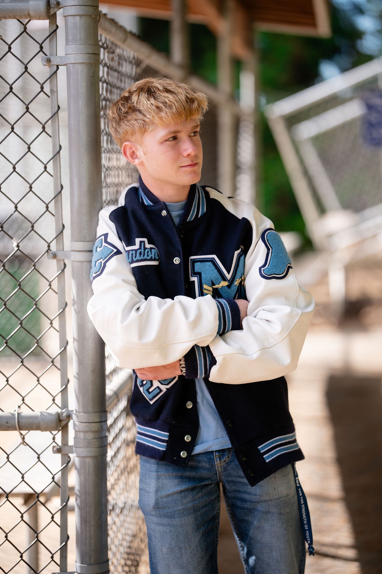 Student in a navy and white varsity jacket and jeans leans against a chain link fence in an outdoor setting.