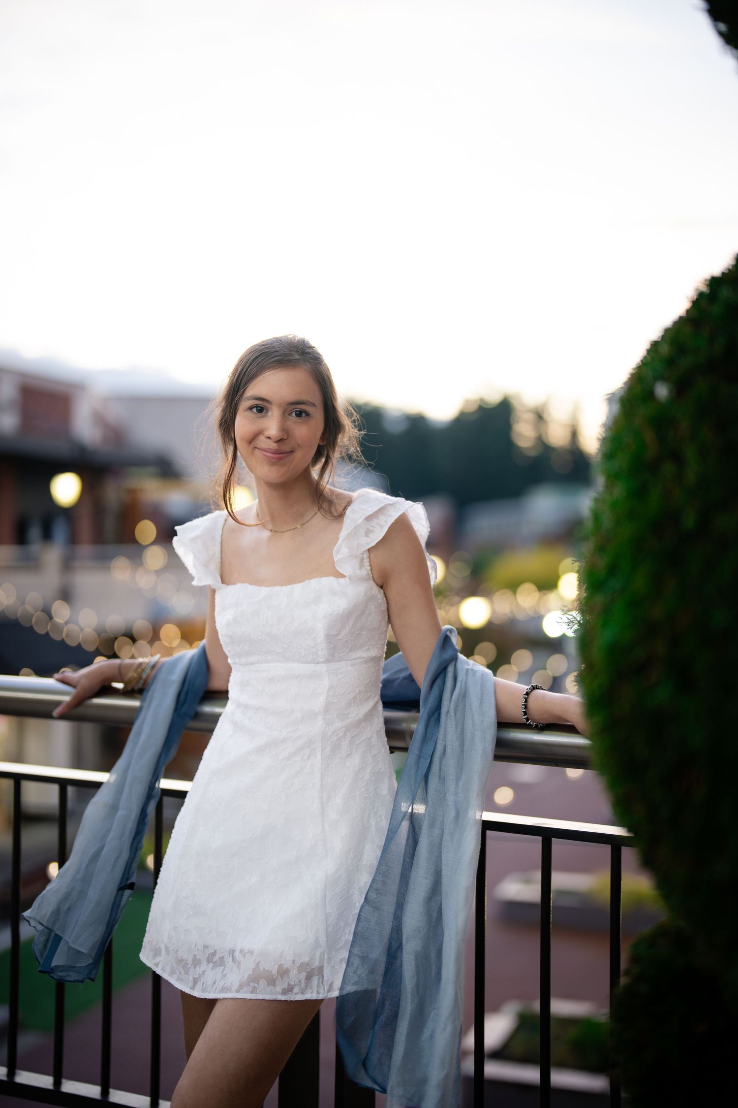 A person in a white summer dress and denim jacket stands on an outdoor balcony at dusk with bokeh lights in background.