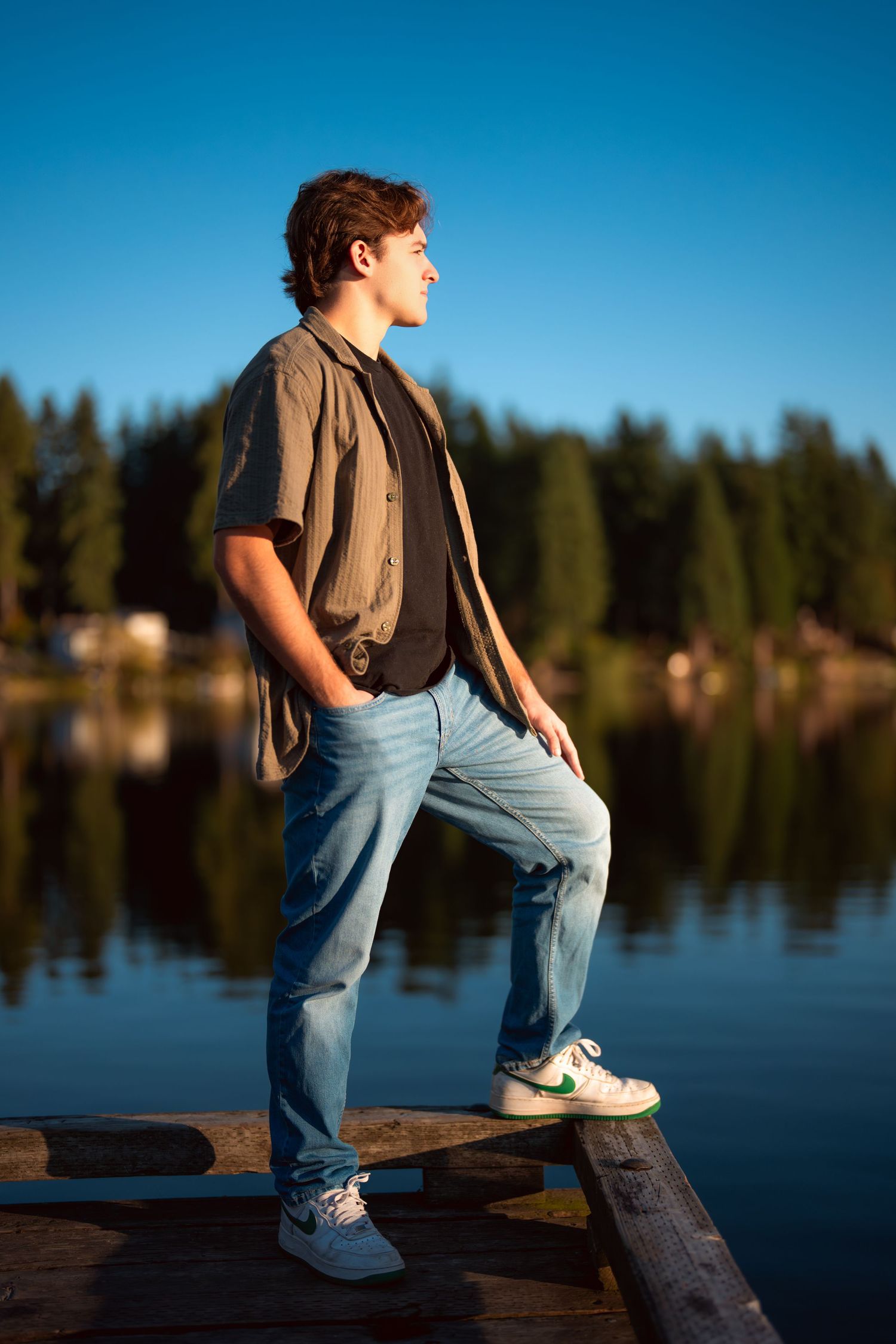 Person in jeans and brown vest sits contemplatively on wooden dock by serene lake during sunset.