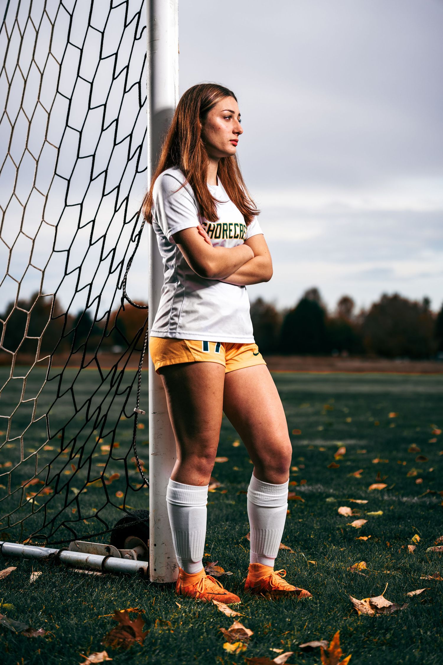 Soccer player in white uniform and orange cleats stands by goal net with autumn leaves scattered on field.