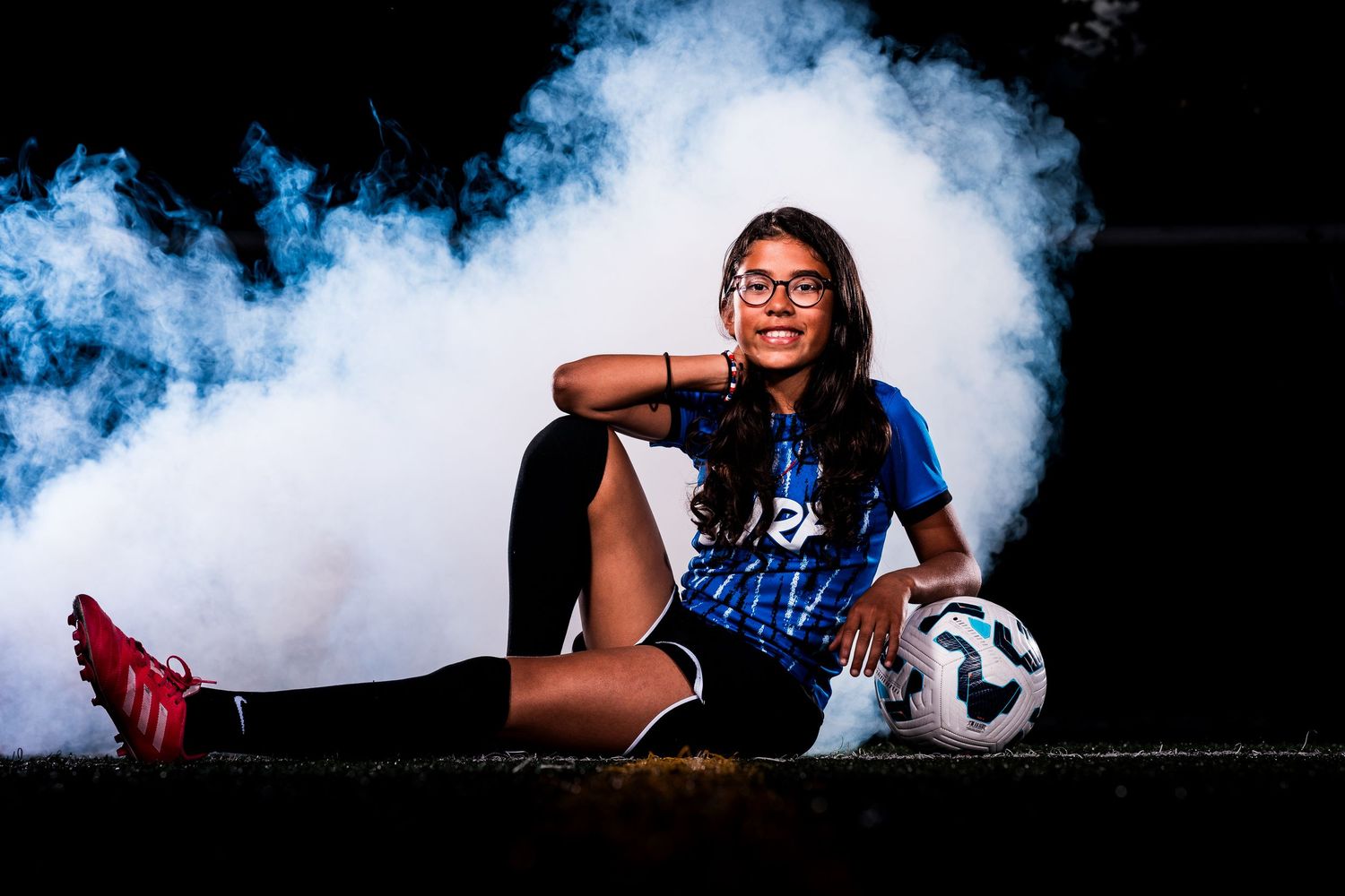 Soccer player poses with ball against dramatic blue smoke background for dynamic sports photography series.