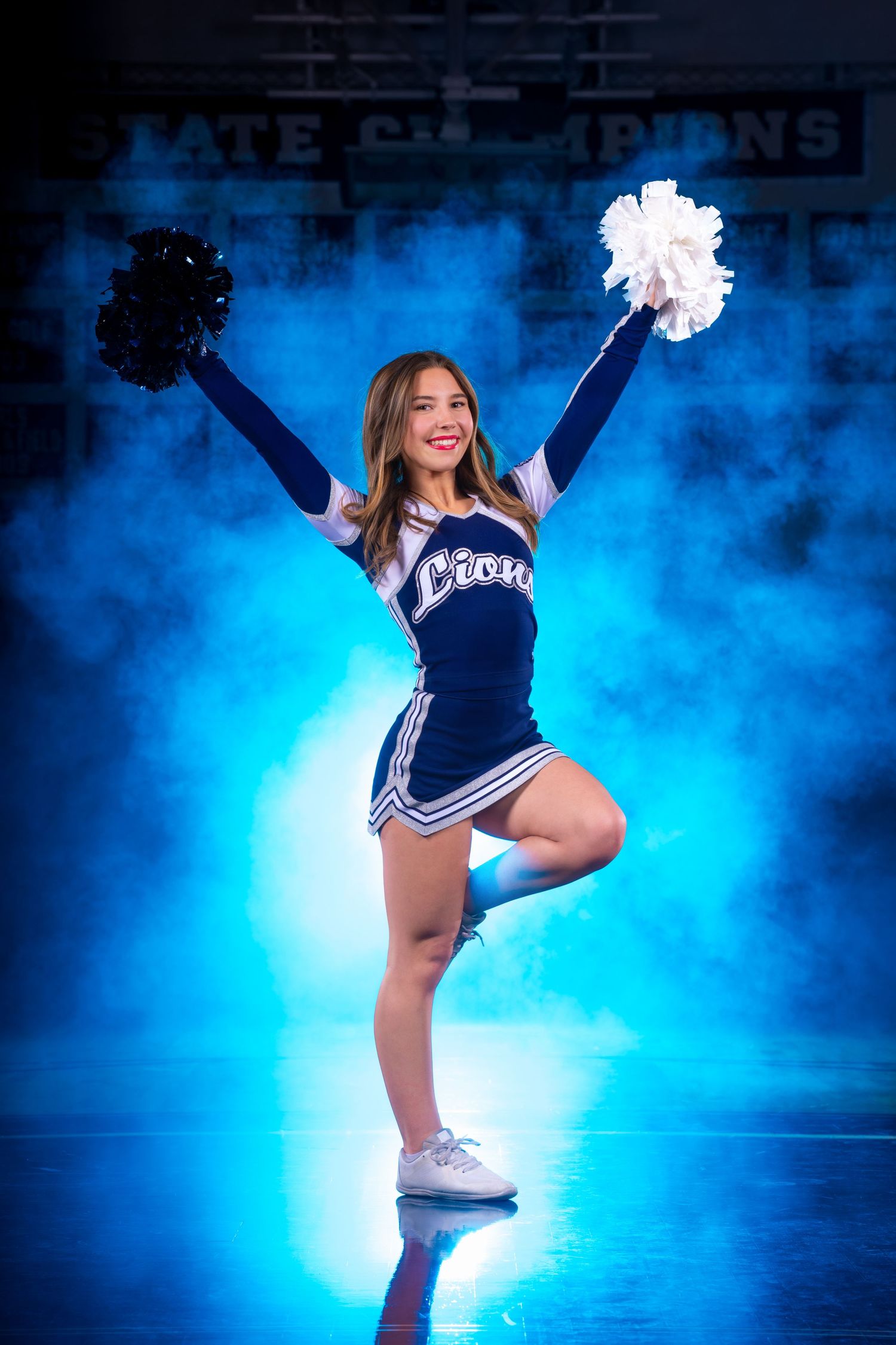 Cheerleader performs high-energy dance routine in navy uniform against dramatic blue smoke-filled backdrop.