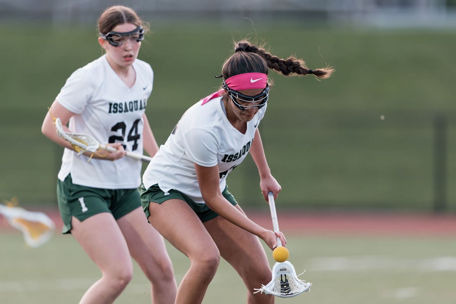 Two lacrosse players compete for possession during a match, with one wearing white and the other in a pink headband.