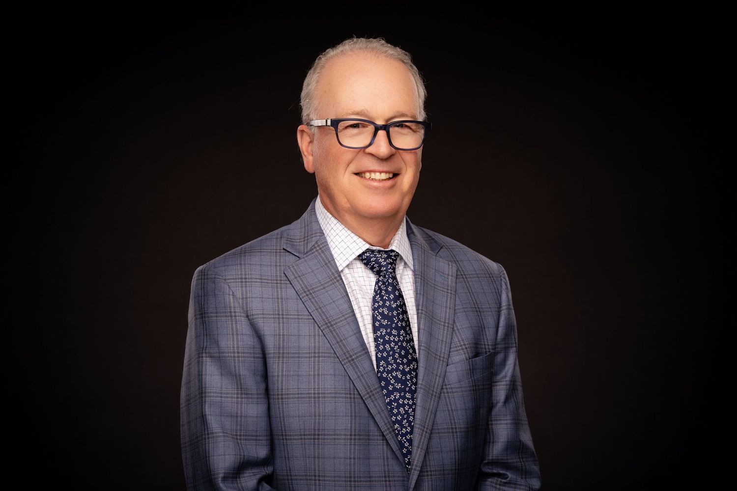 Professional headshot of person wearing gray plaid suit jacket with polka dot tie and glasses against dark background.