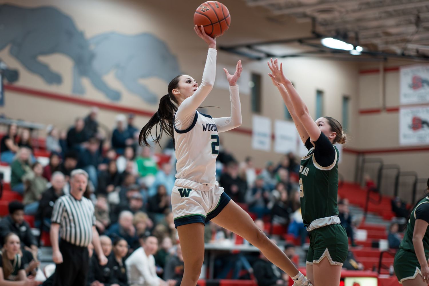 Basketball player leaps high to make a shot during an intense game between teams in white and green uniforms.