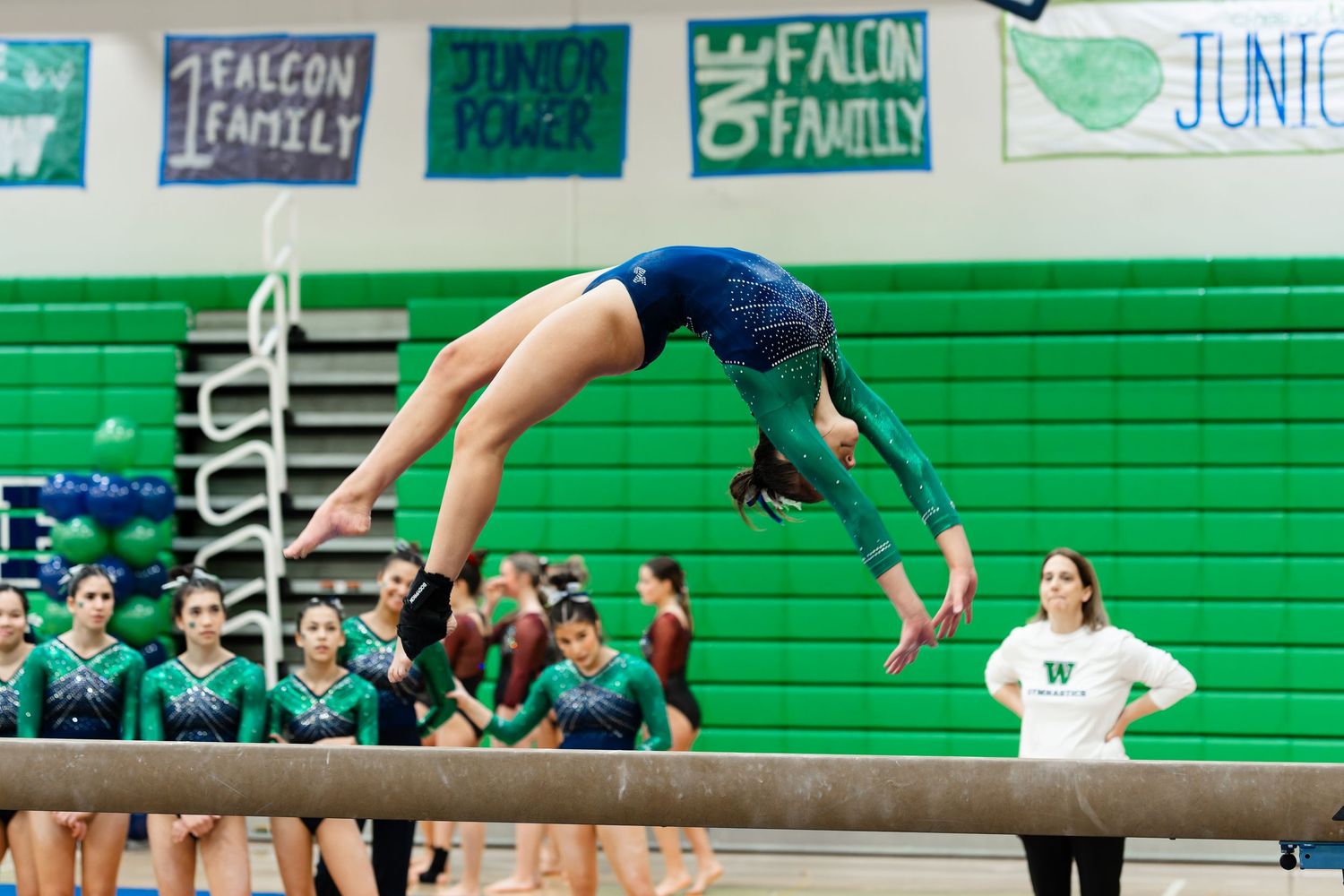 High school gymnast performs an impressive balance beam routine during competition in a green-lit gymnasium.