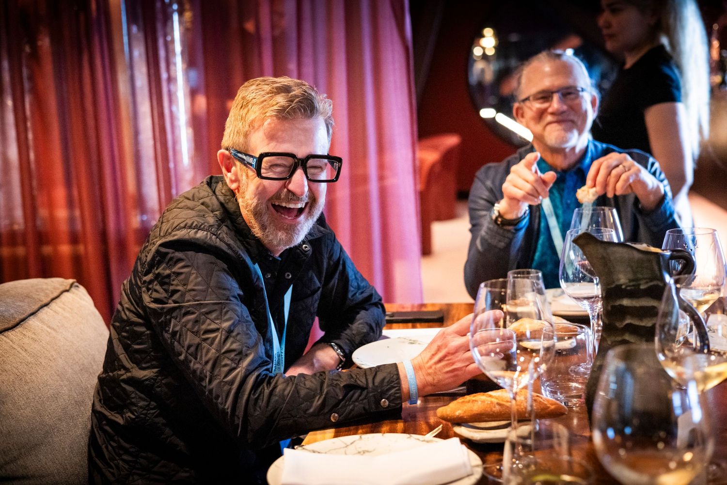 Man in black jacket and bold glasses laughing joyfully during a formal dinner event.