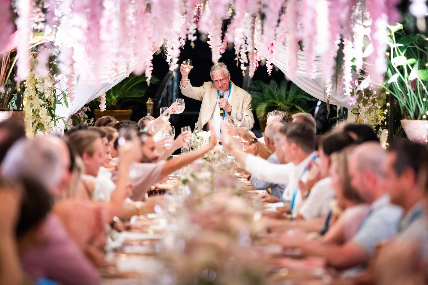 Festive wedding banquet with guests toasting as an animated speaker stands at a decorated long table.