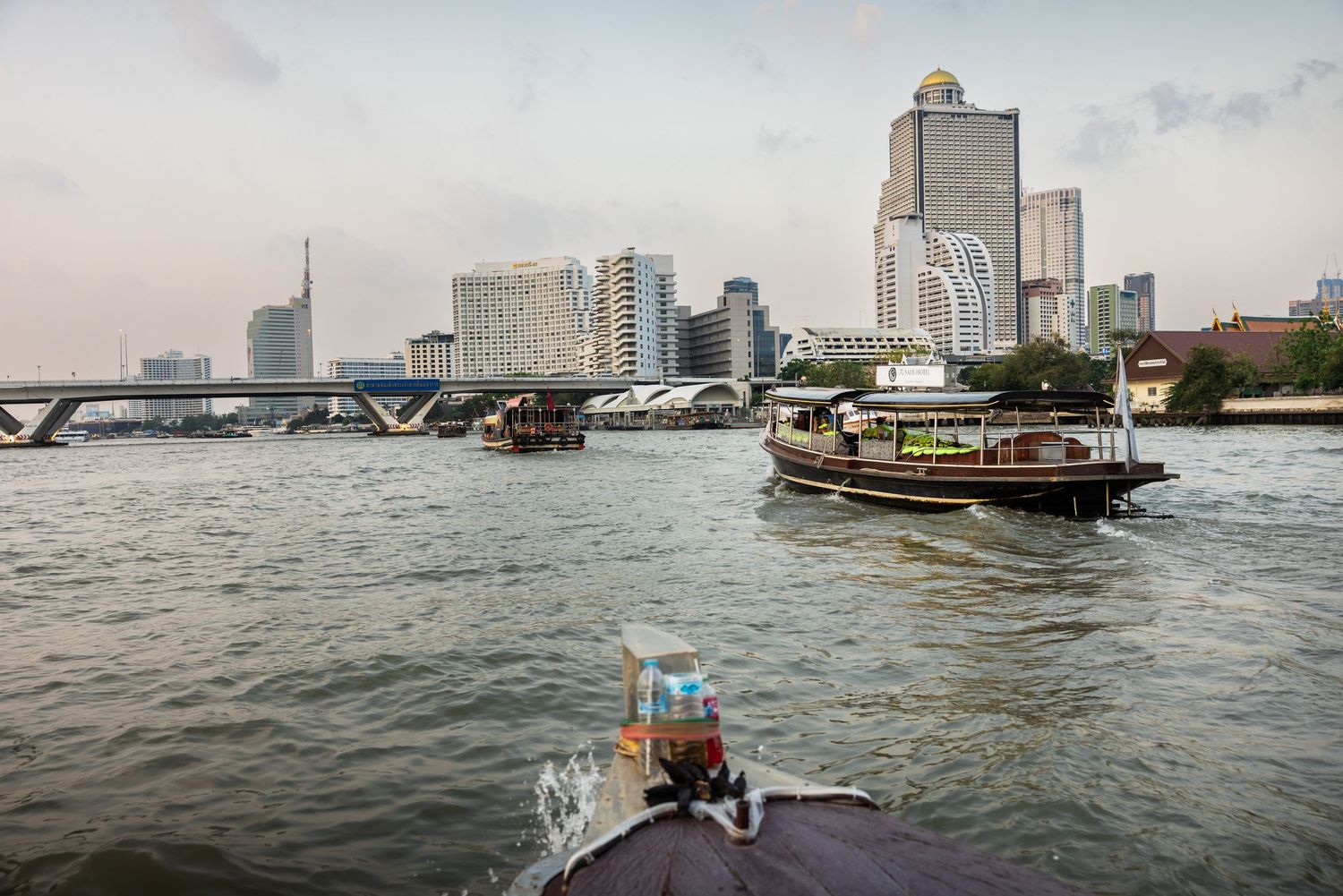 Boats navigating a busy urban river with city skyline at dusk in Bangkok.