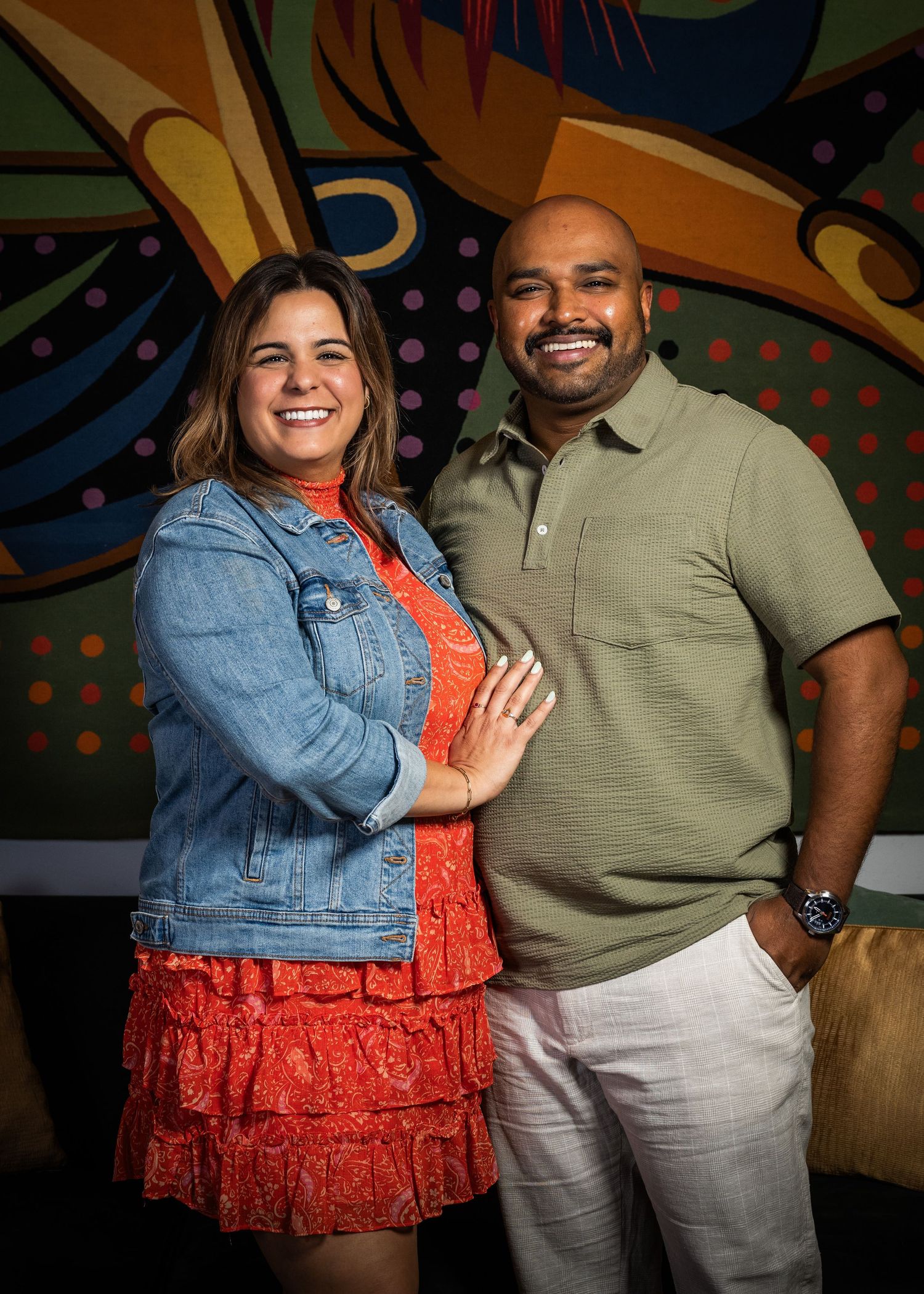 A smiling couple poses together in front of a colorful mural at a casual venue.