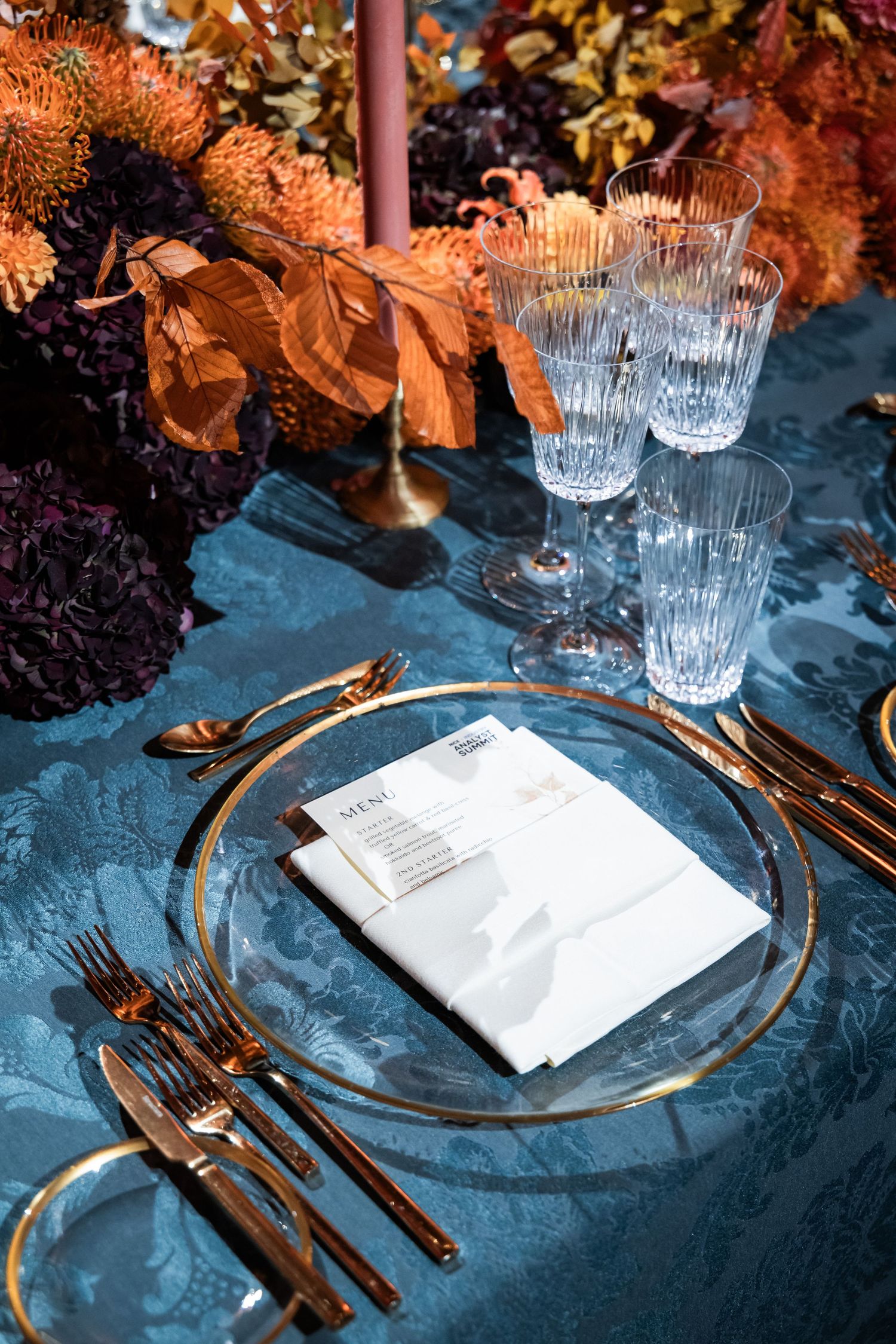 Elegant formal place setting with crystal glasses, gold cutlery, and autumn floral centerpiece on blue tablecloth.