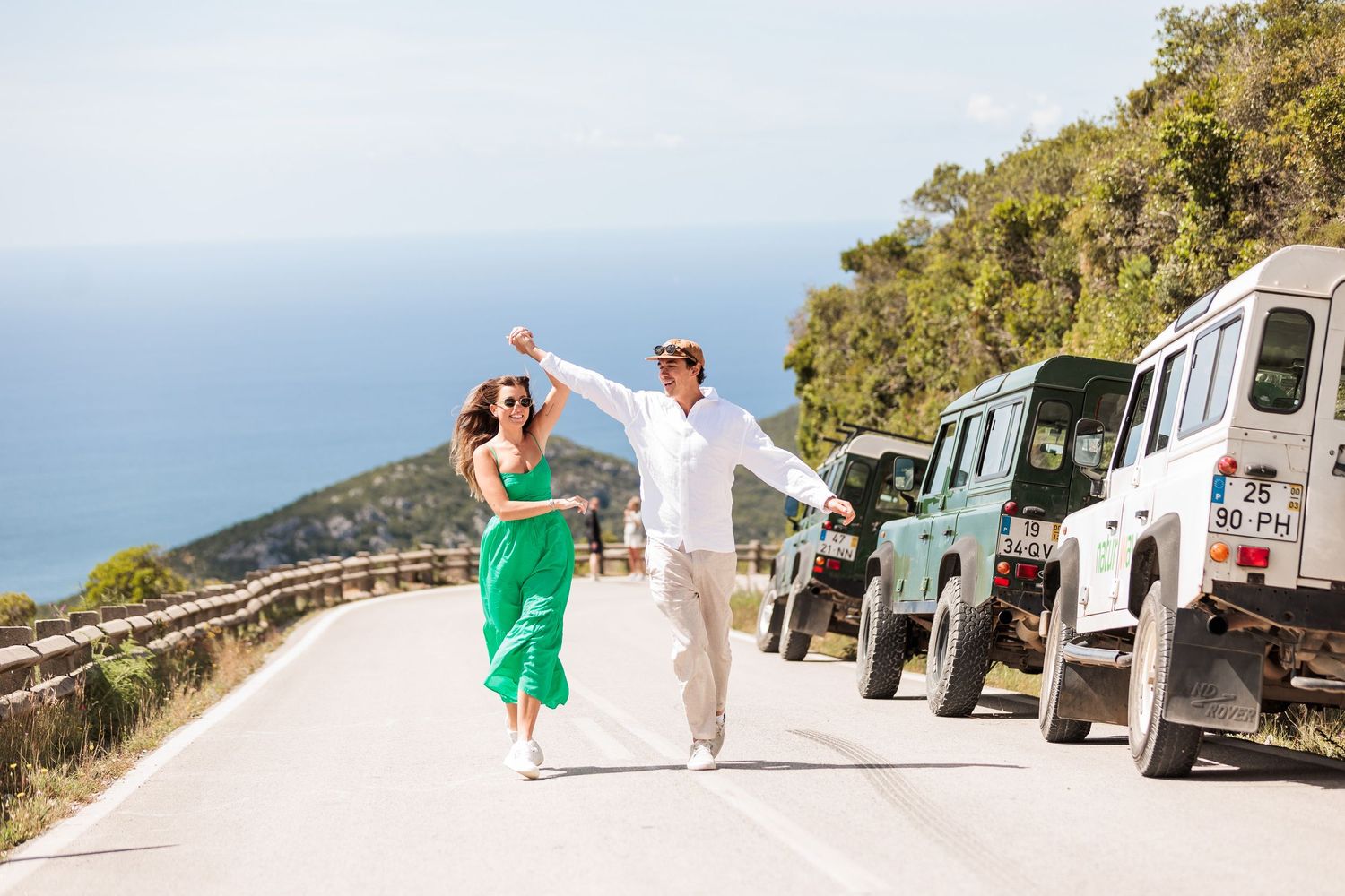 Couple in elegant outfits joyfully walking on a coastal road with Land Rover Defenders and blue sea behind.