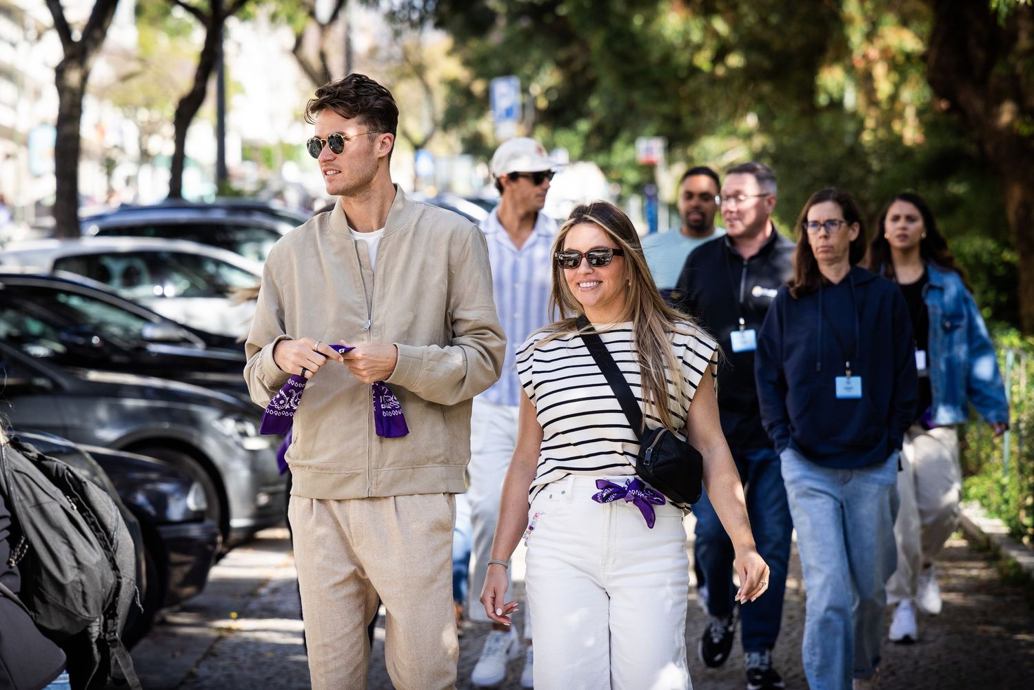 A stylish couple walks hand in hand along a sunny tree-lined street at an outdoor event.