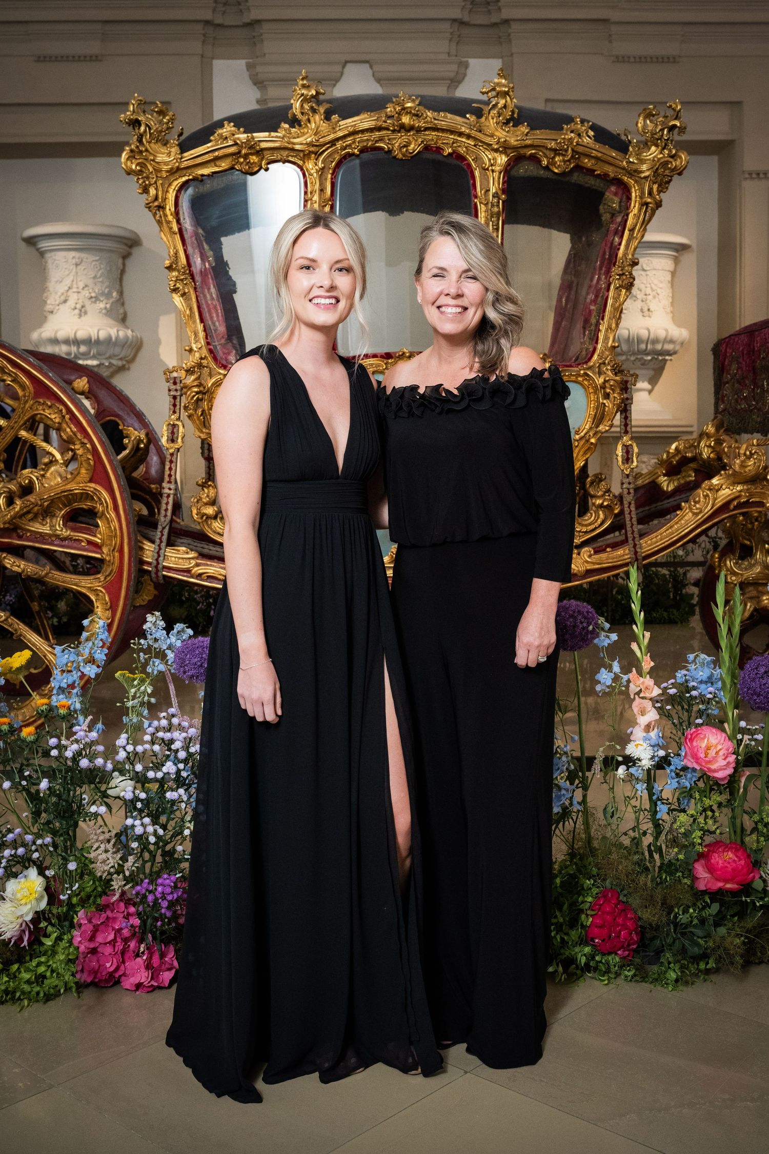 Two women in elegant black gowns pose before an ornate gold carriage surrounded by colorful flowers.