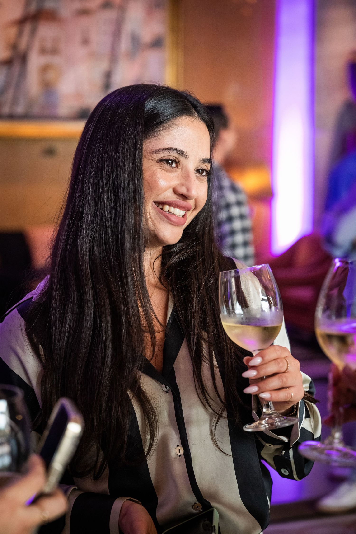 Smiling dark-haired woman holding a glass of white wine at a vibrant evening social gathering.