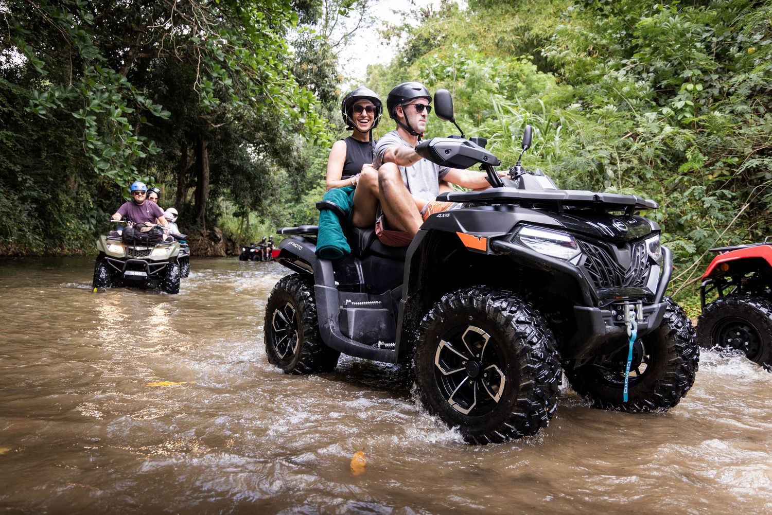 Riders on ATVs crossing a shallow river stream through a lush tropical jungle trail.
