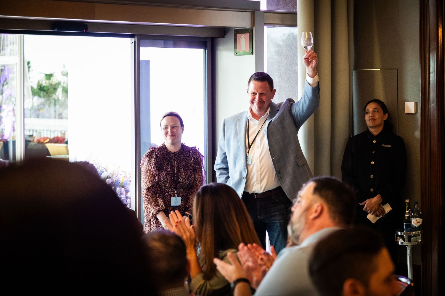 A man raises his glass triumphantly while guests applaud at an indoor celebration event.