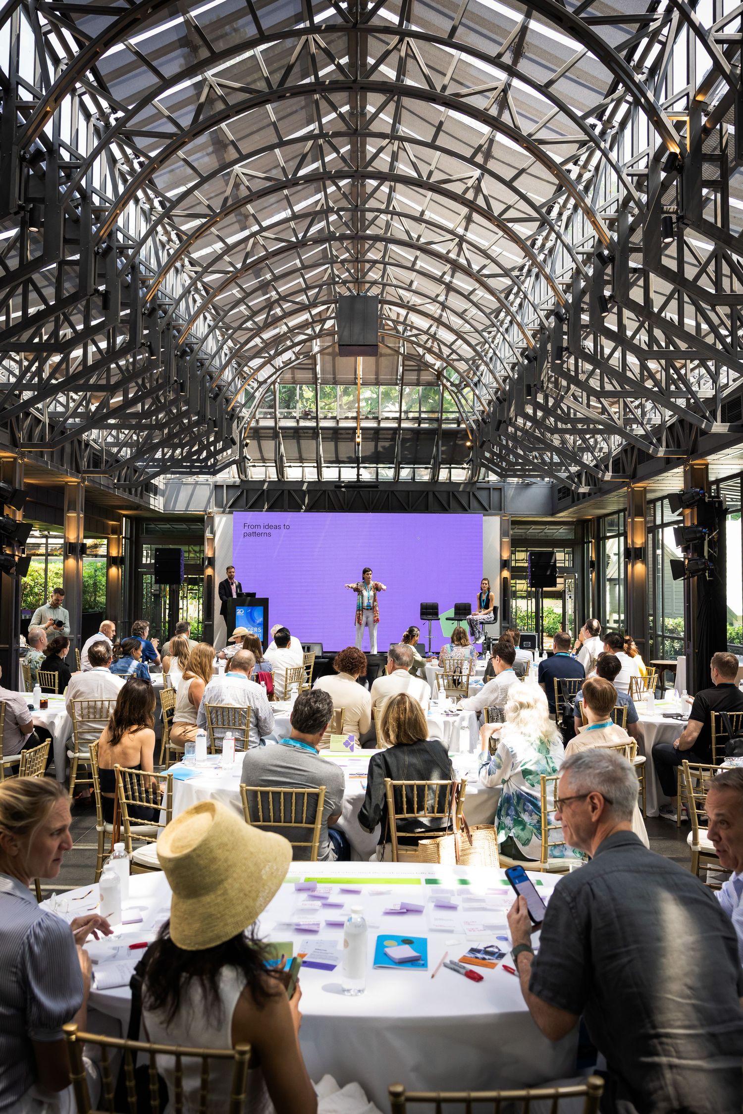 Large indoor event venue with glass ceiling, purple stage screen, and seated audience at round tables.