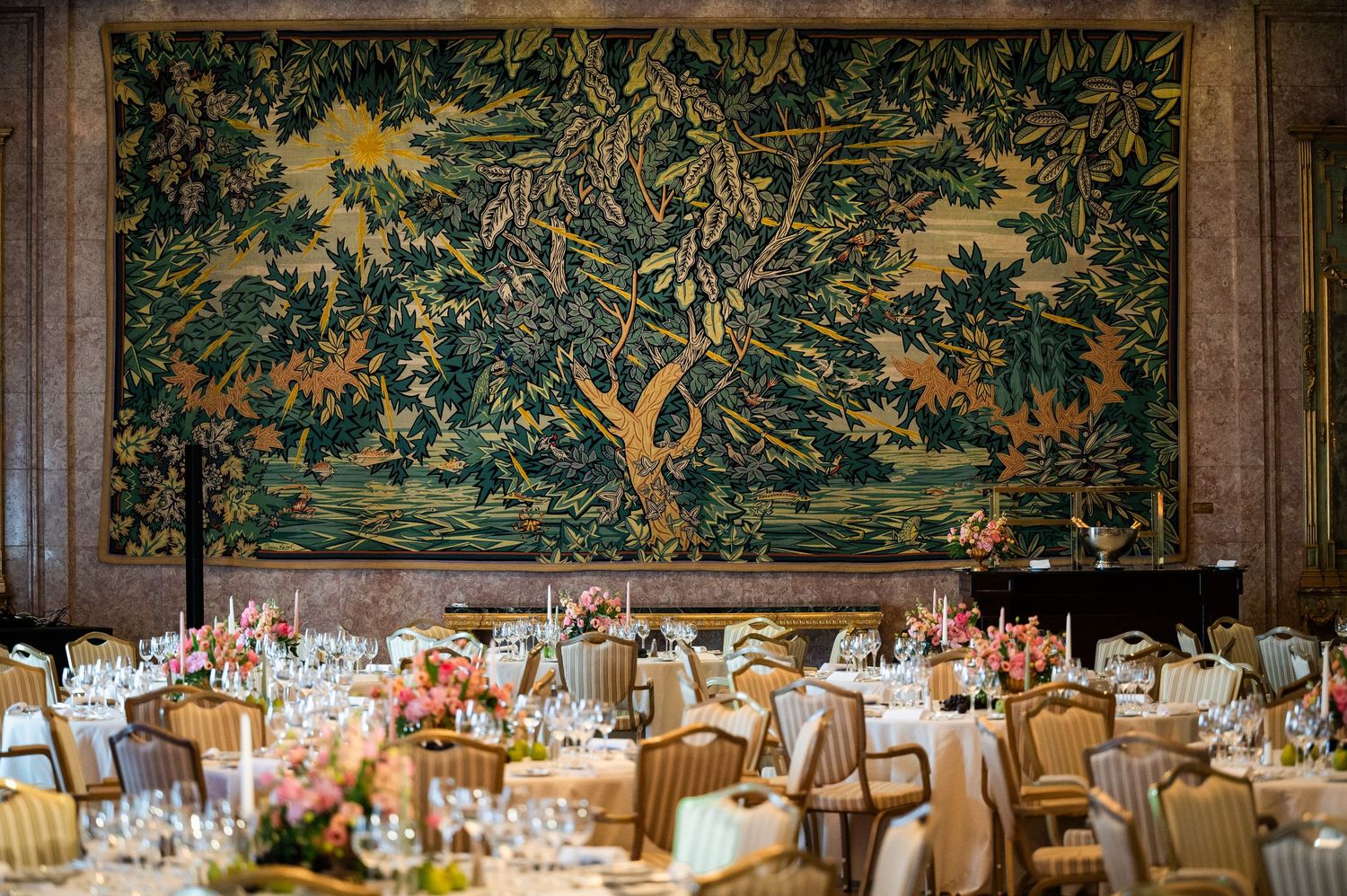 Elegant banquet hall with round tables set for dinner beneath a large ornate tapestry wall hanging.