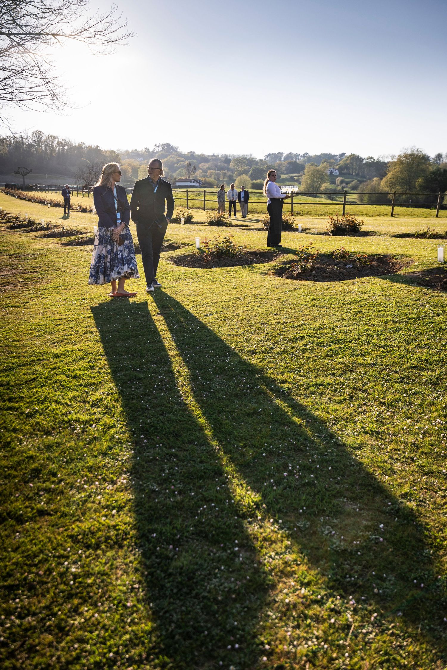 Two people walking through a sunlit garden casting long shadows during an outdoor conference event.