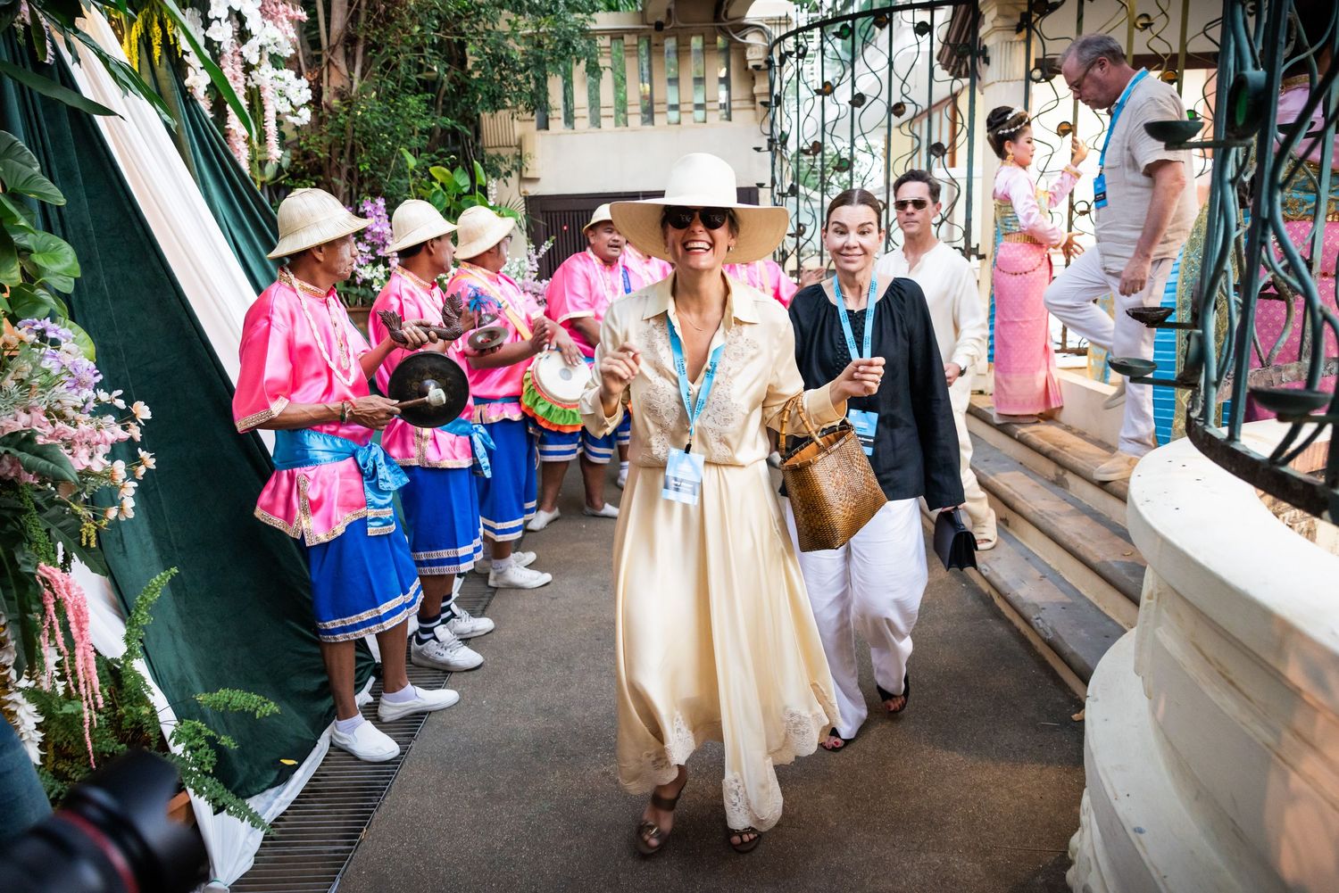 Woman in cream dress and white hat leads group past Thai musicians in pink traditional costumes.