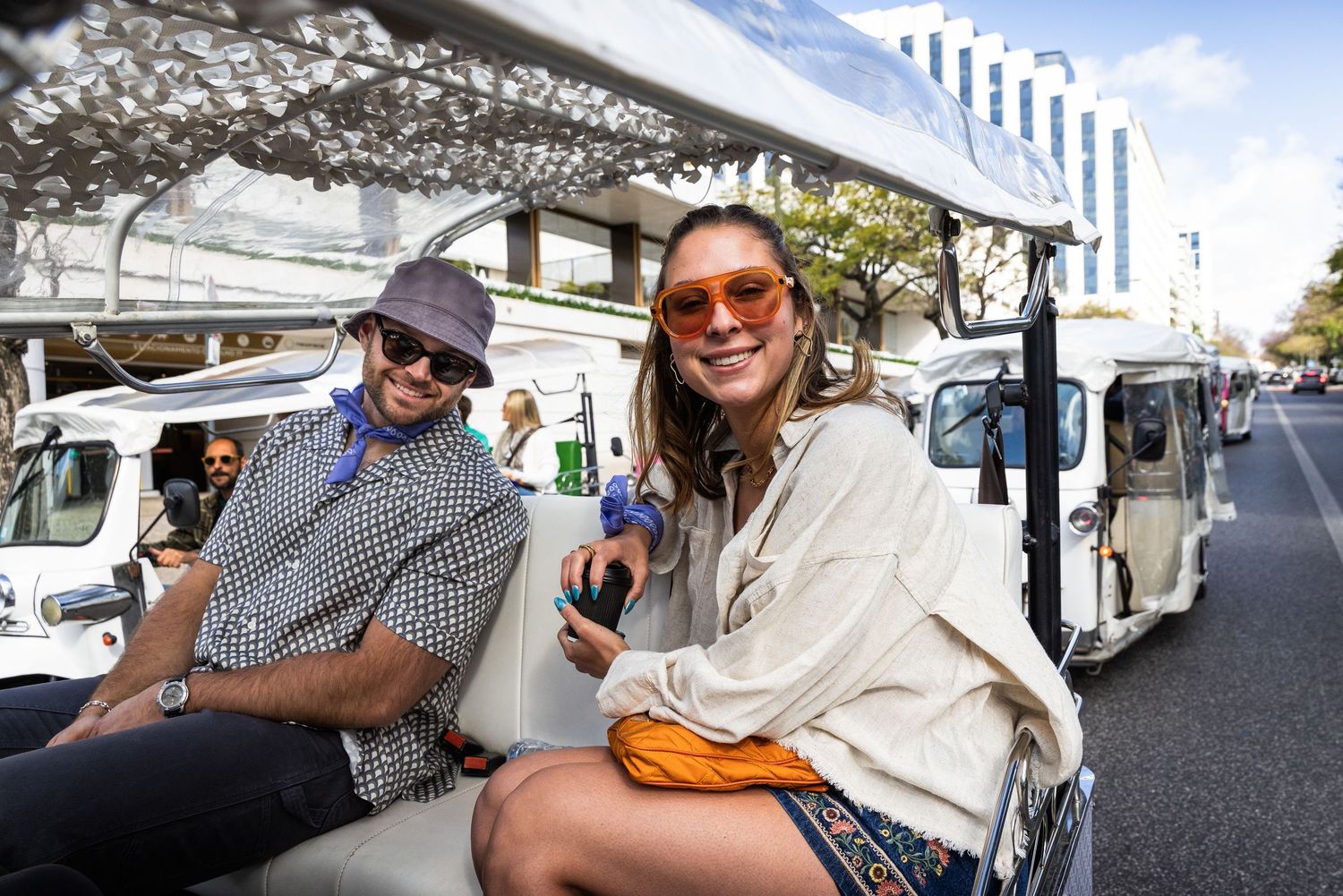 A smiling couple wearing sunglasses enjoys a tuk-tuk sightseeing tour on a sunny day.