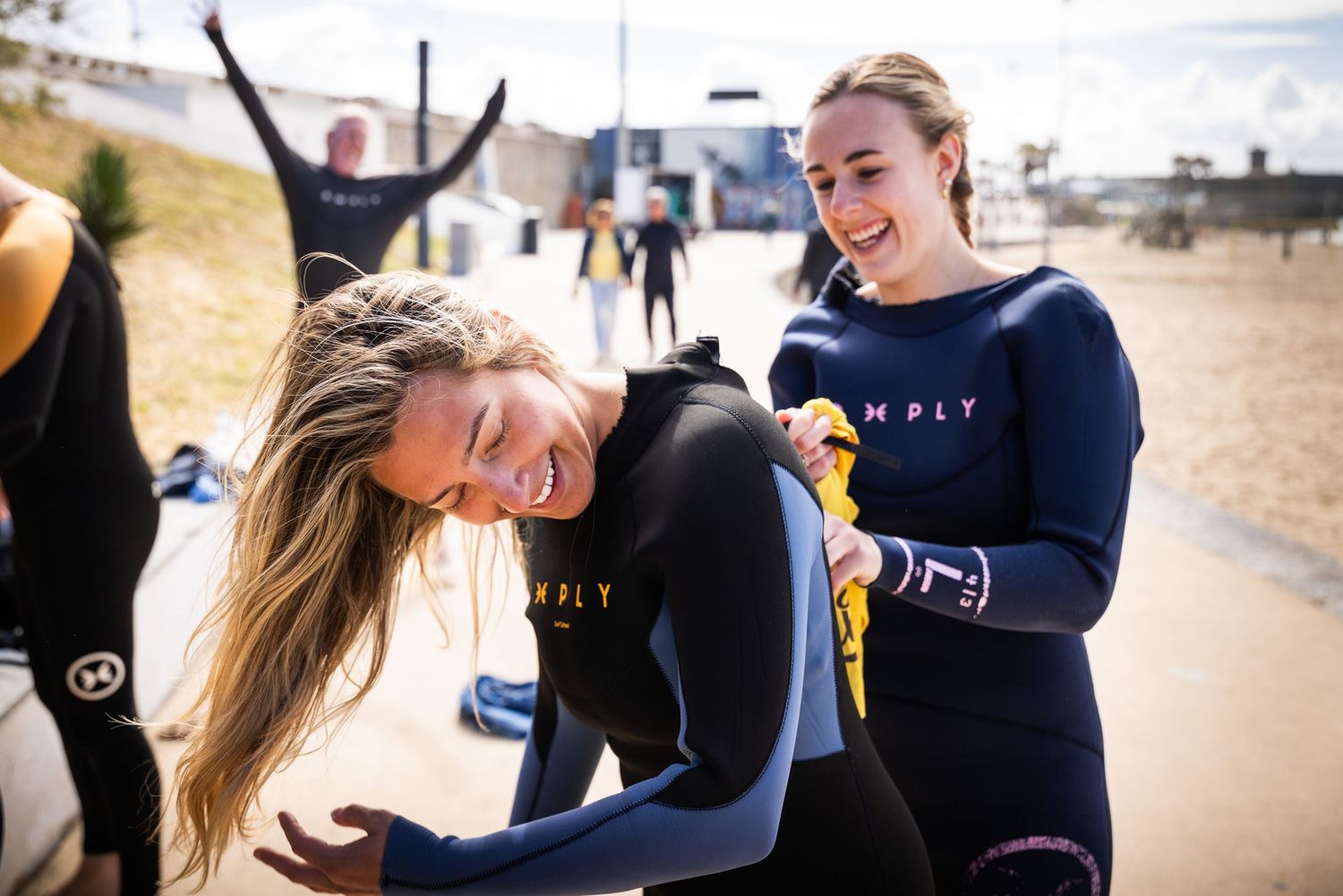 Two smiling women in HMPLY wetsuits helping each other prepare for a surf lesson on the beach.