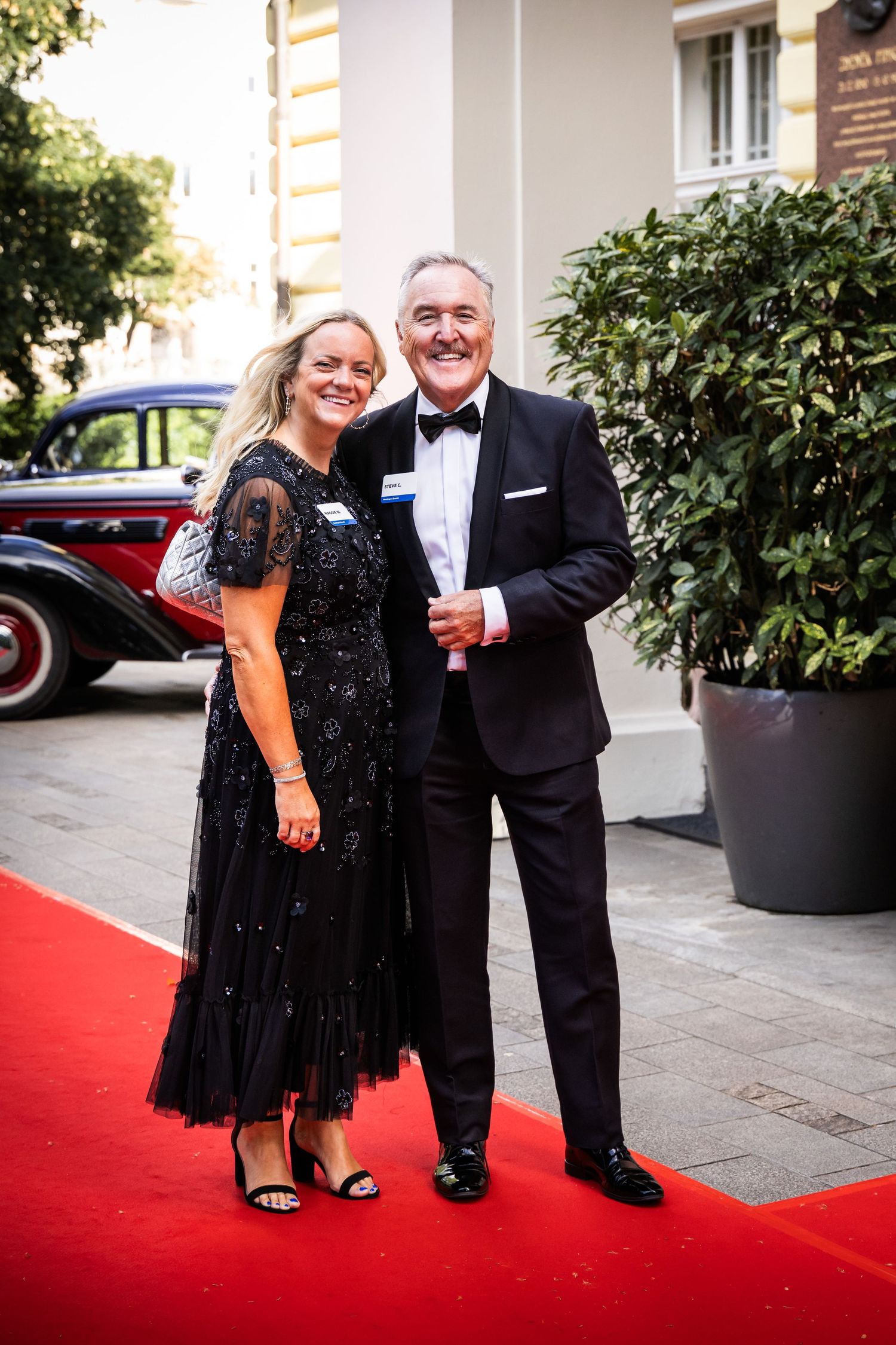 Couple posing on a red carpet at a formal black-tie outdoor evening event.