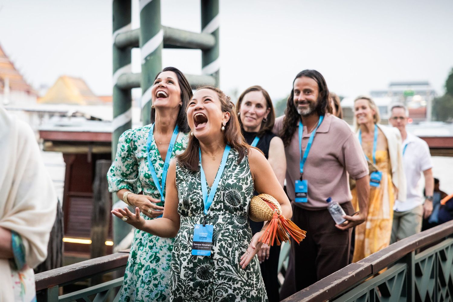 Group of joyful conference attendees laughing and reacting outdoors wearing blue lanyards near a waterway.