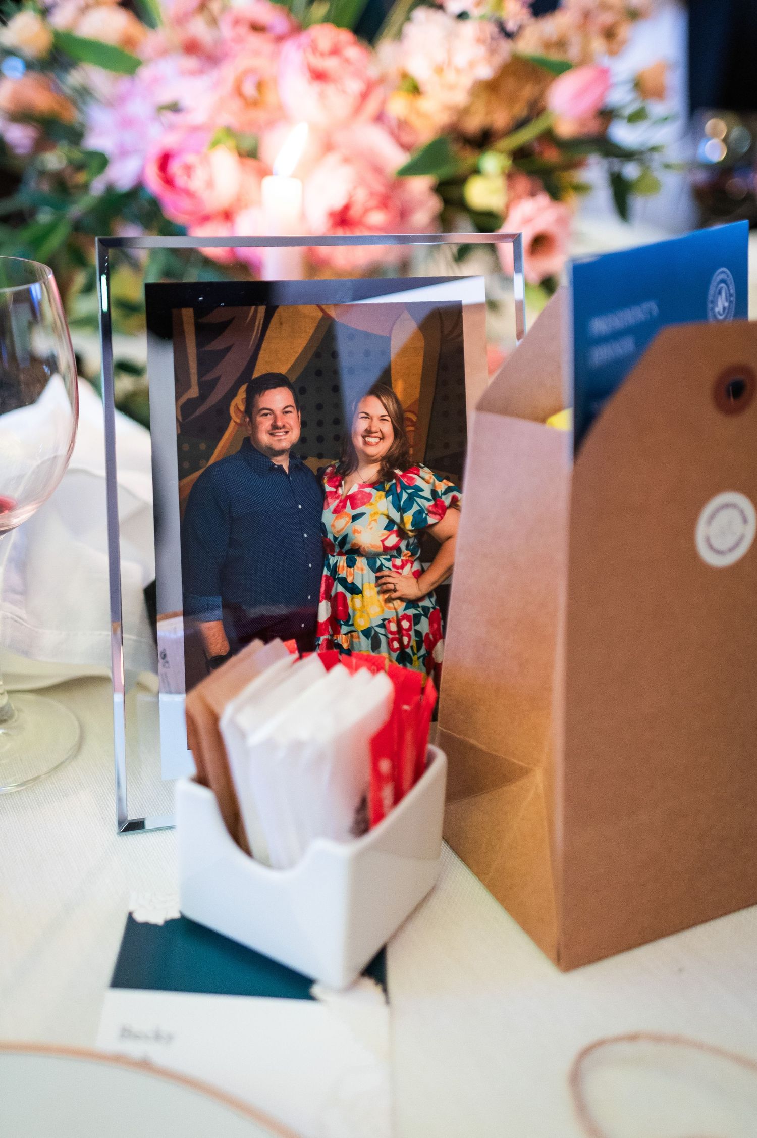 A framed photo of a smiling couple displayed on a wedding table with pink floral centerpiece and gift bags.