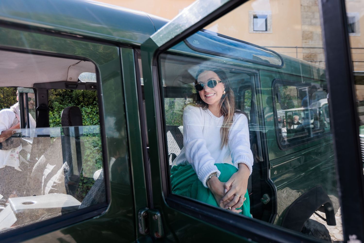 Smiling woman in sunglasses and green skirt sitting in open door of vintage green Land Rover Defender.