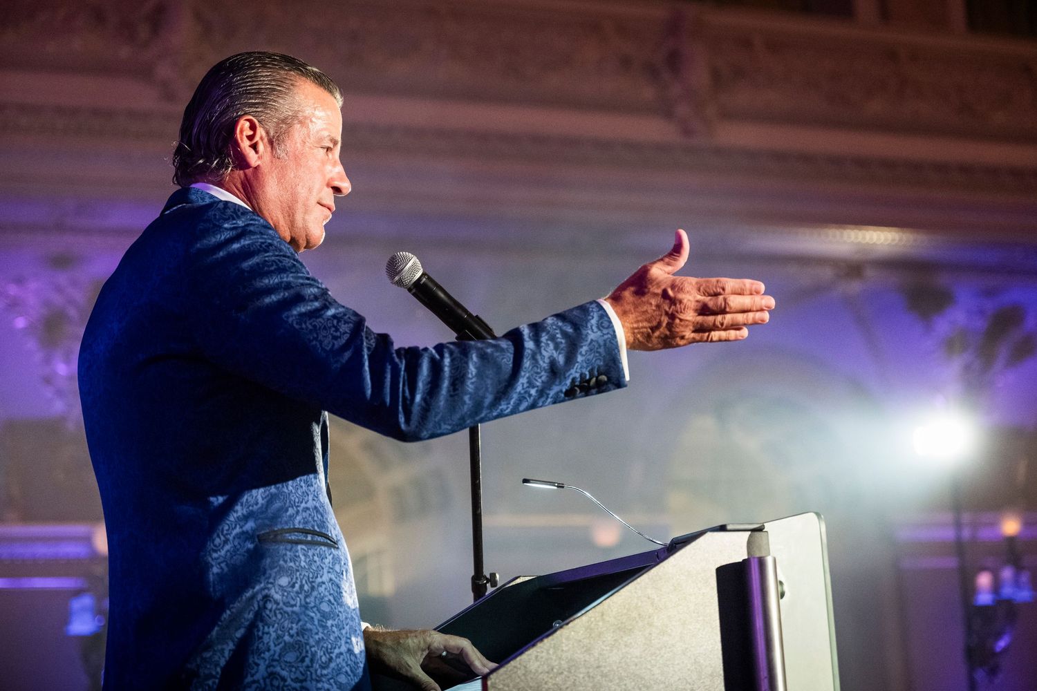 Dynamic speaker in blue blazer gesturing at podium with microphone during formal evening event.