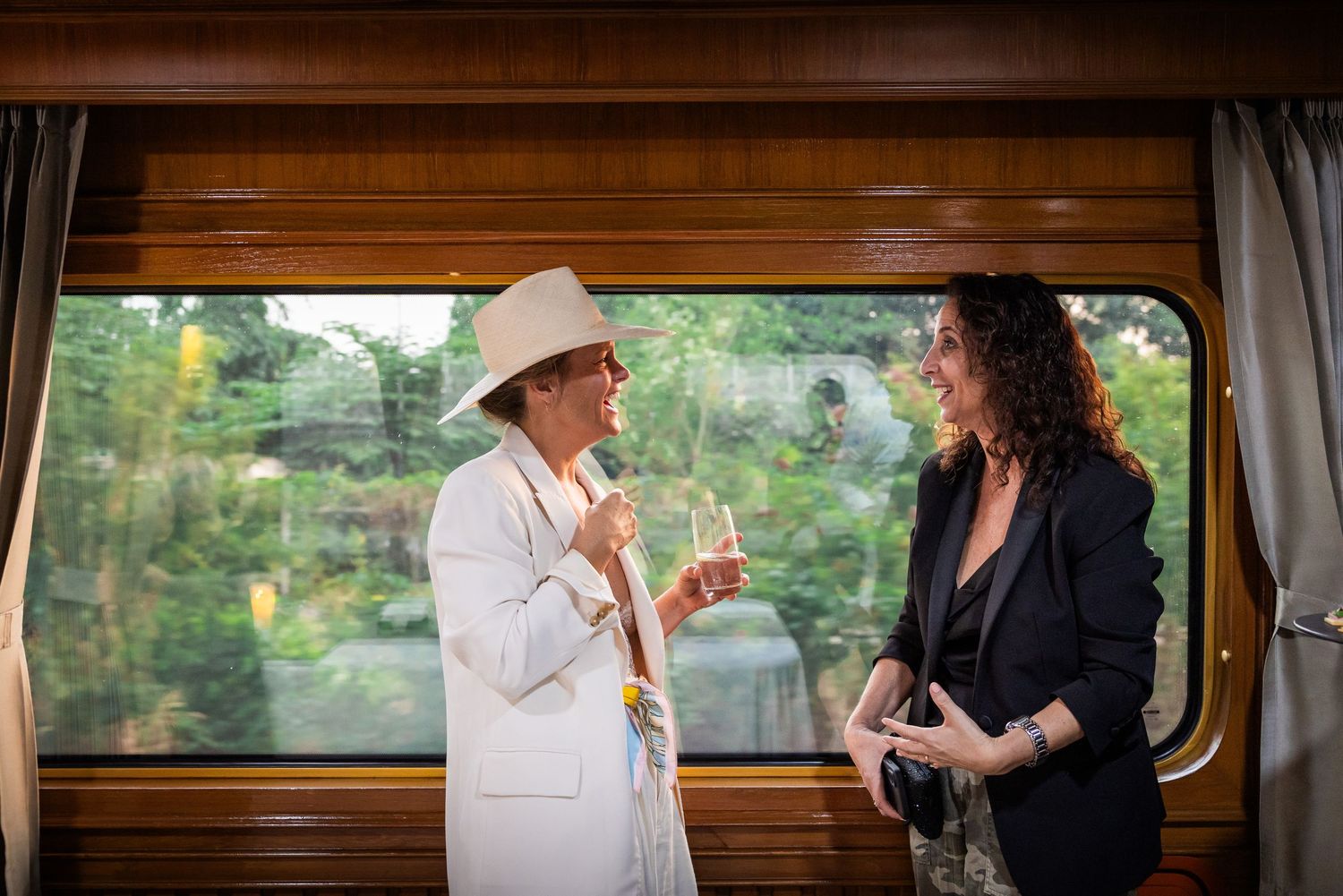 Two elegantly dressed guests chatting with champagne by a window on a luxury train.