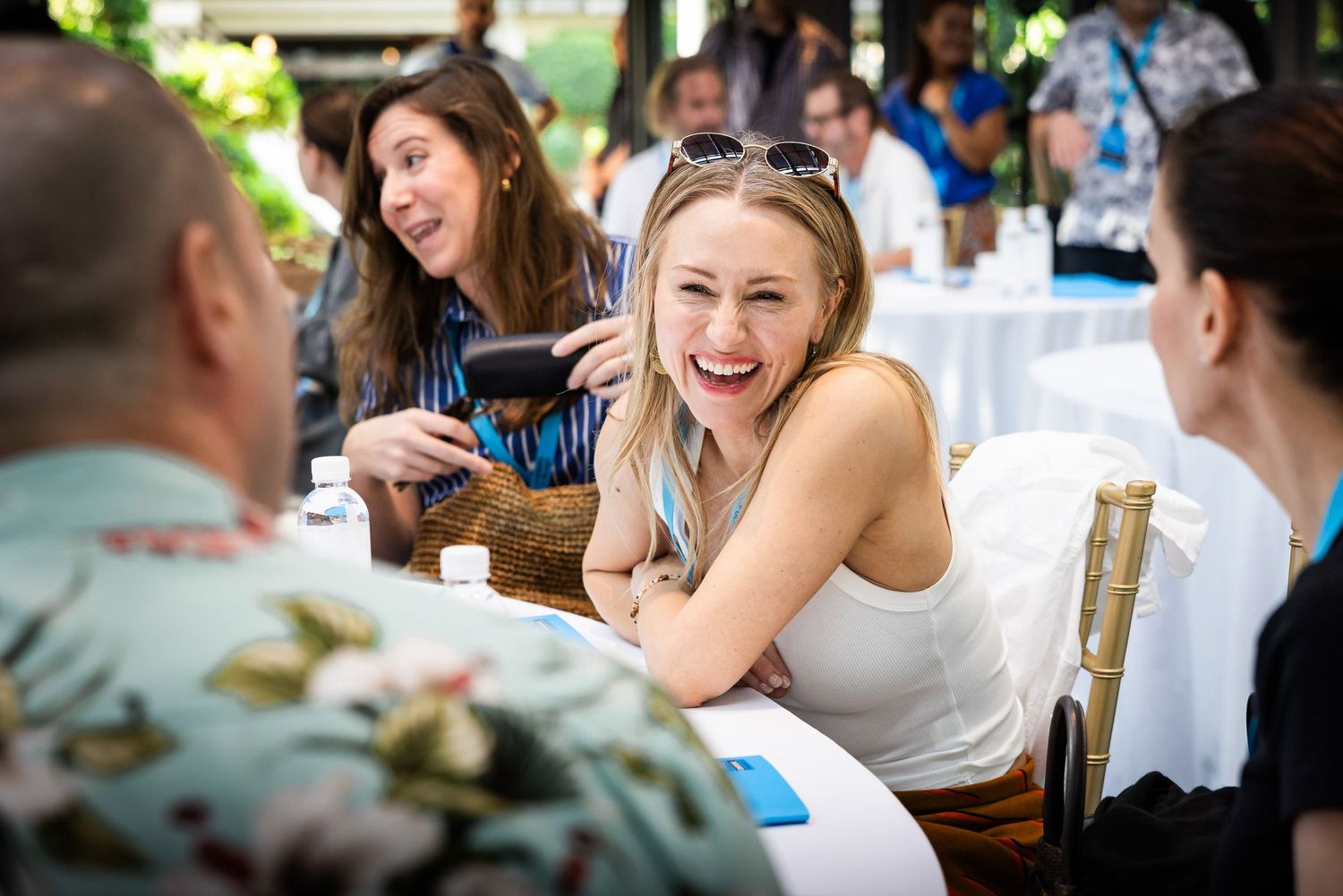 Young blonde woman laughing joyfully at an outdoor dining event with other guests at white tables.