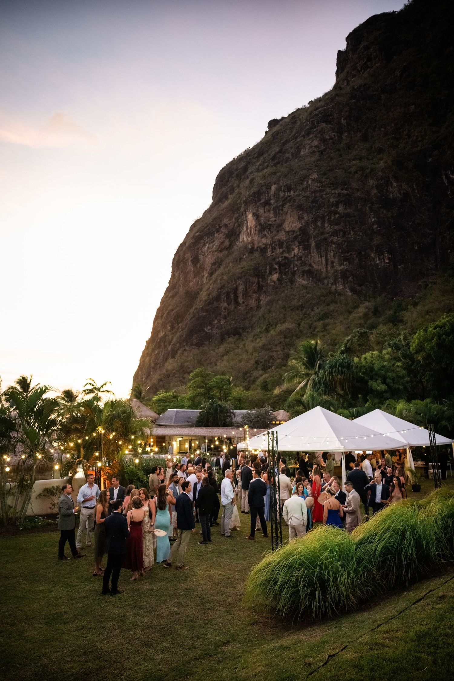Outdoor evening event with guests mingling beneath string lights at the base of a dramatic cliff.