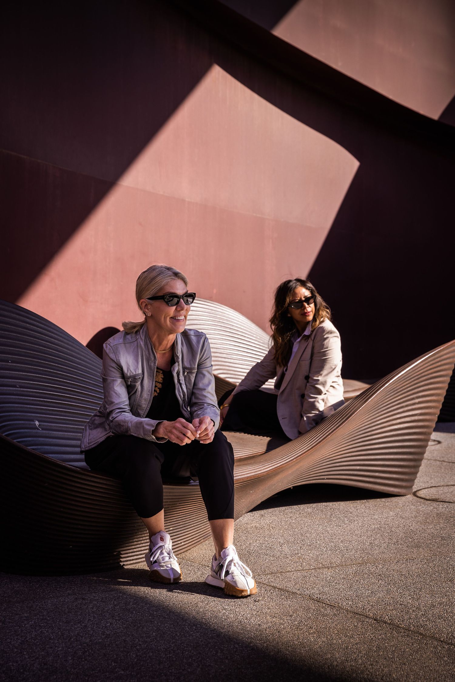 Two women in sunglasses and matching jackets relax on modern curved benches against a red wall.