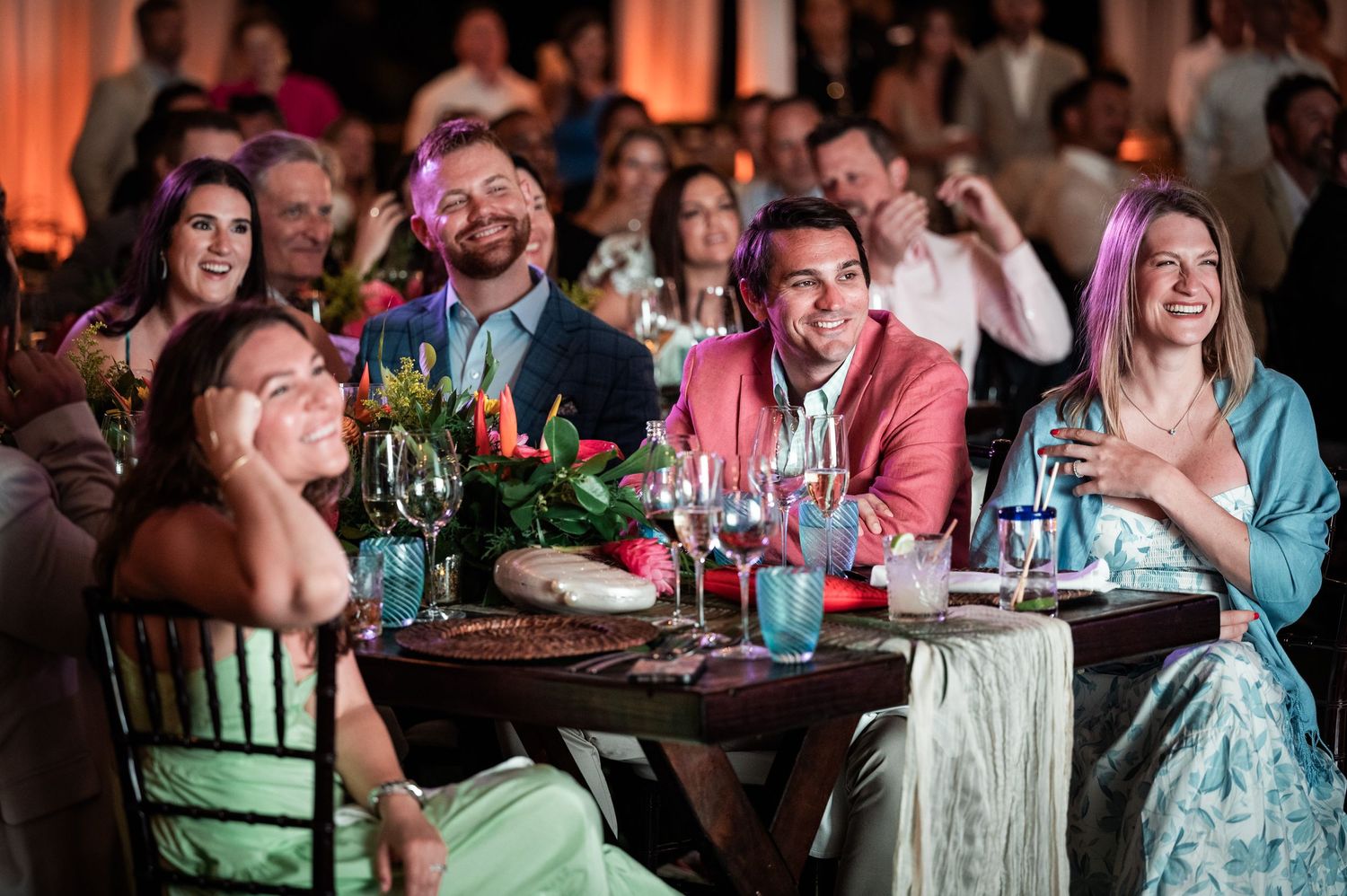 Guests laughing and celebrating at a festive dinner event with tropical floral centerpieces and colorful lighting.