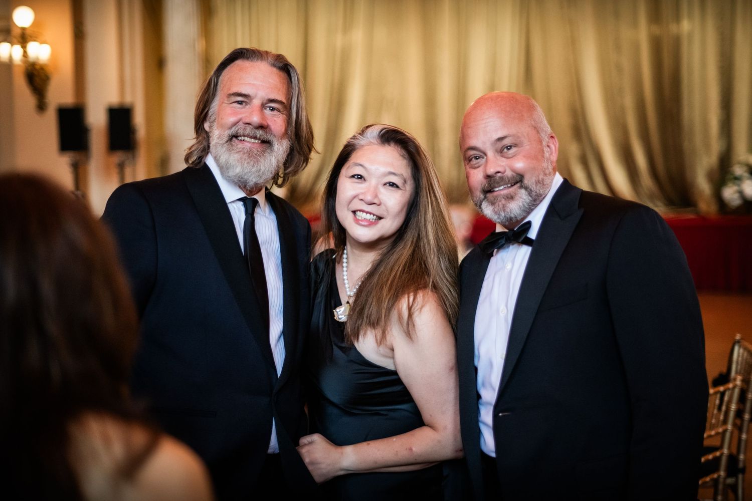 Three people smiling together at an elegant formal gala event in a decorated venue.